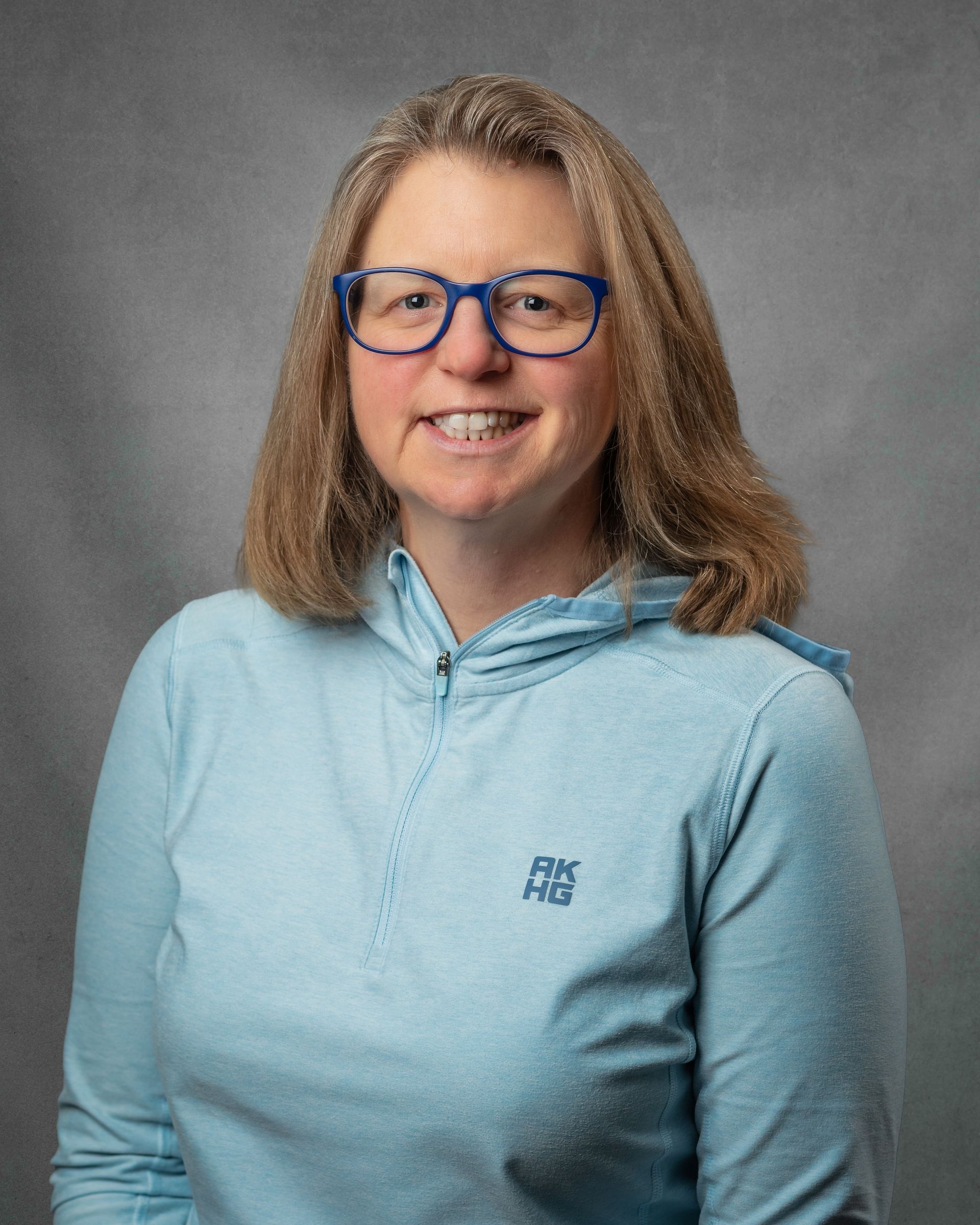 A woman with blue glasses and a nlue shirt smiling in front of a gray background.