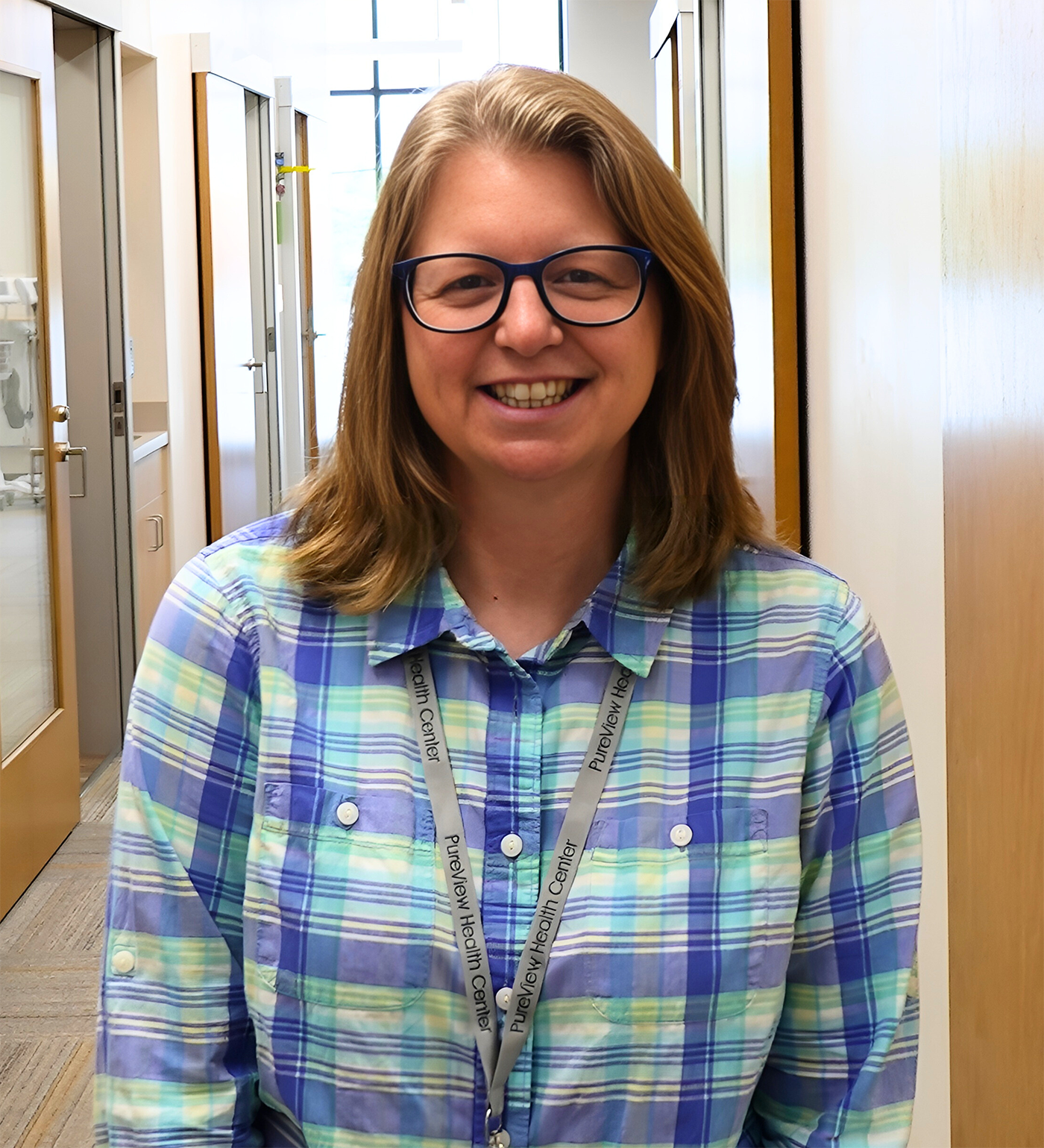 A woman wearing glasses and a plaid shirt is smiling in a hallway.