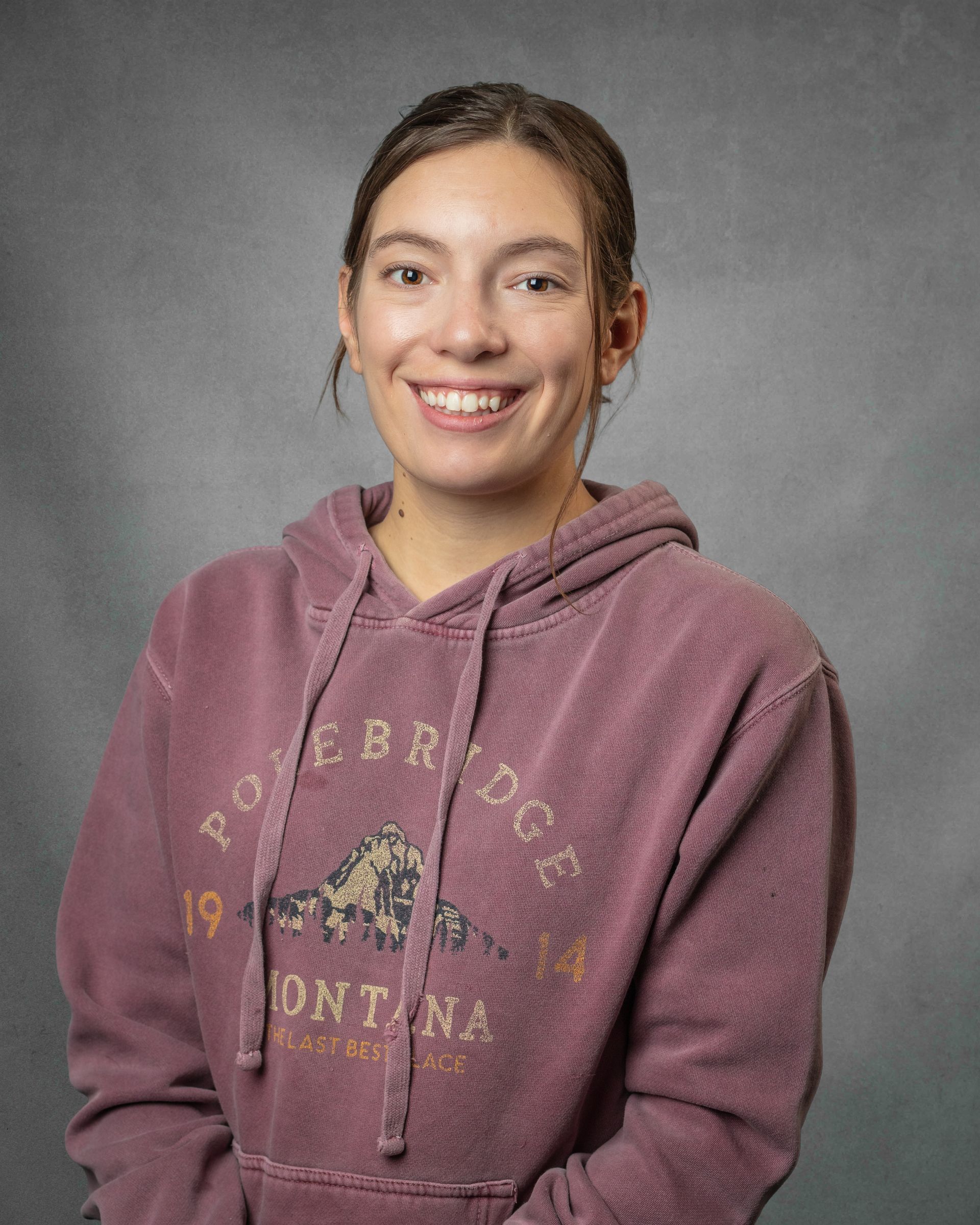 A woman wearing a faded red hoodie is smiling in front of a gray background.