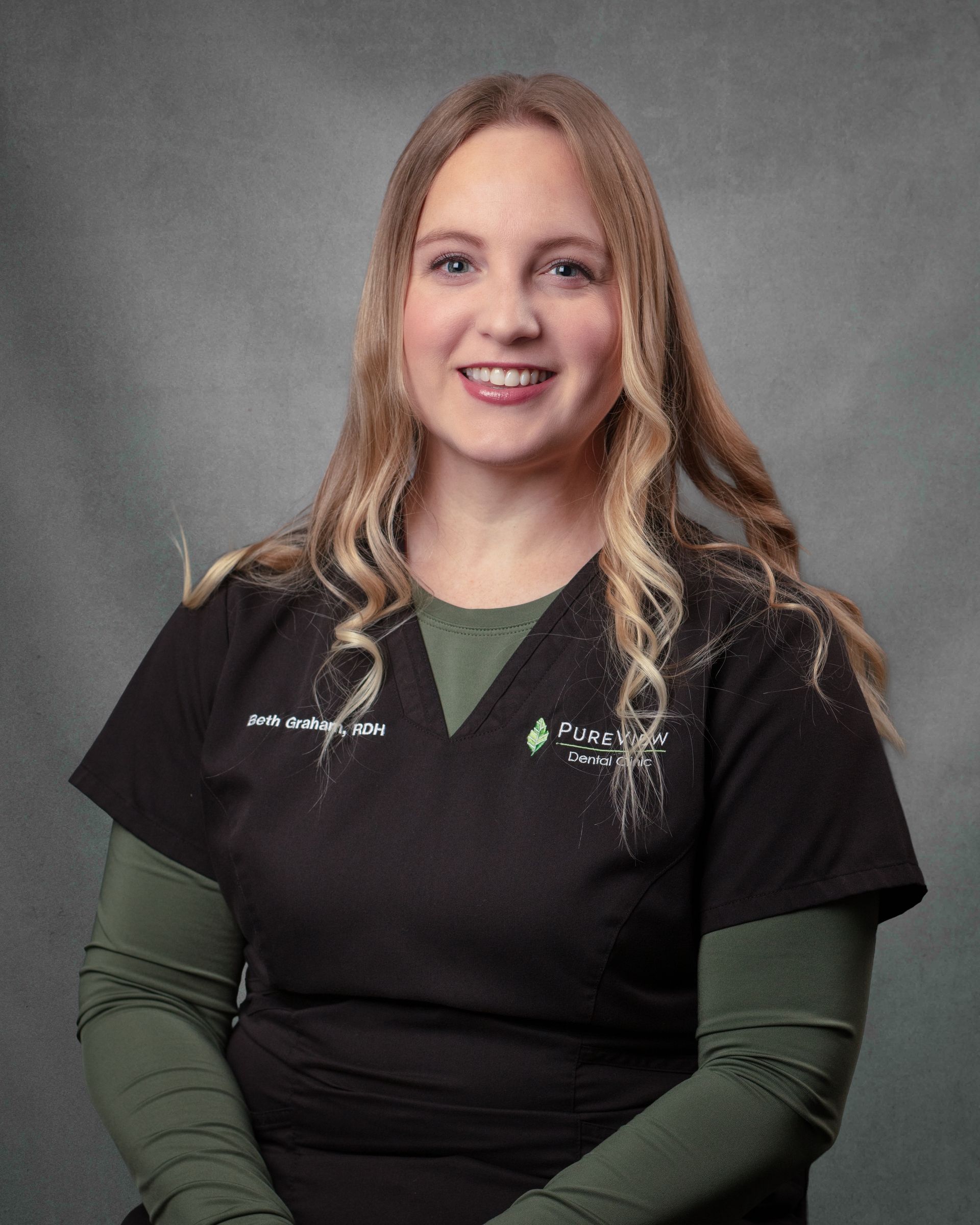 A woman in black scrubs is smiling in front of a gray background.