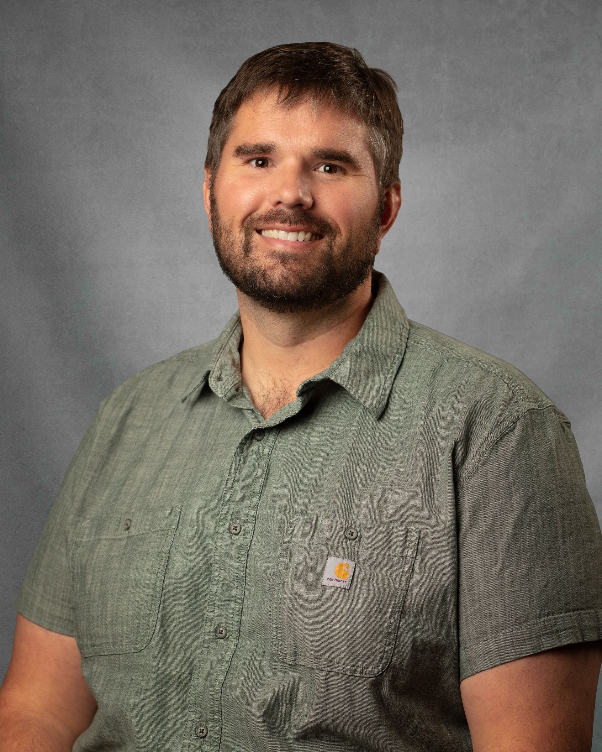 A man with a beard in a green shirt is smiling in front of a gray background.