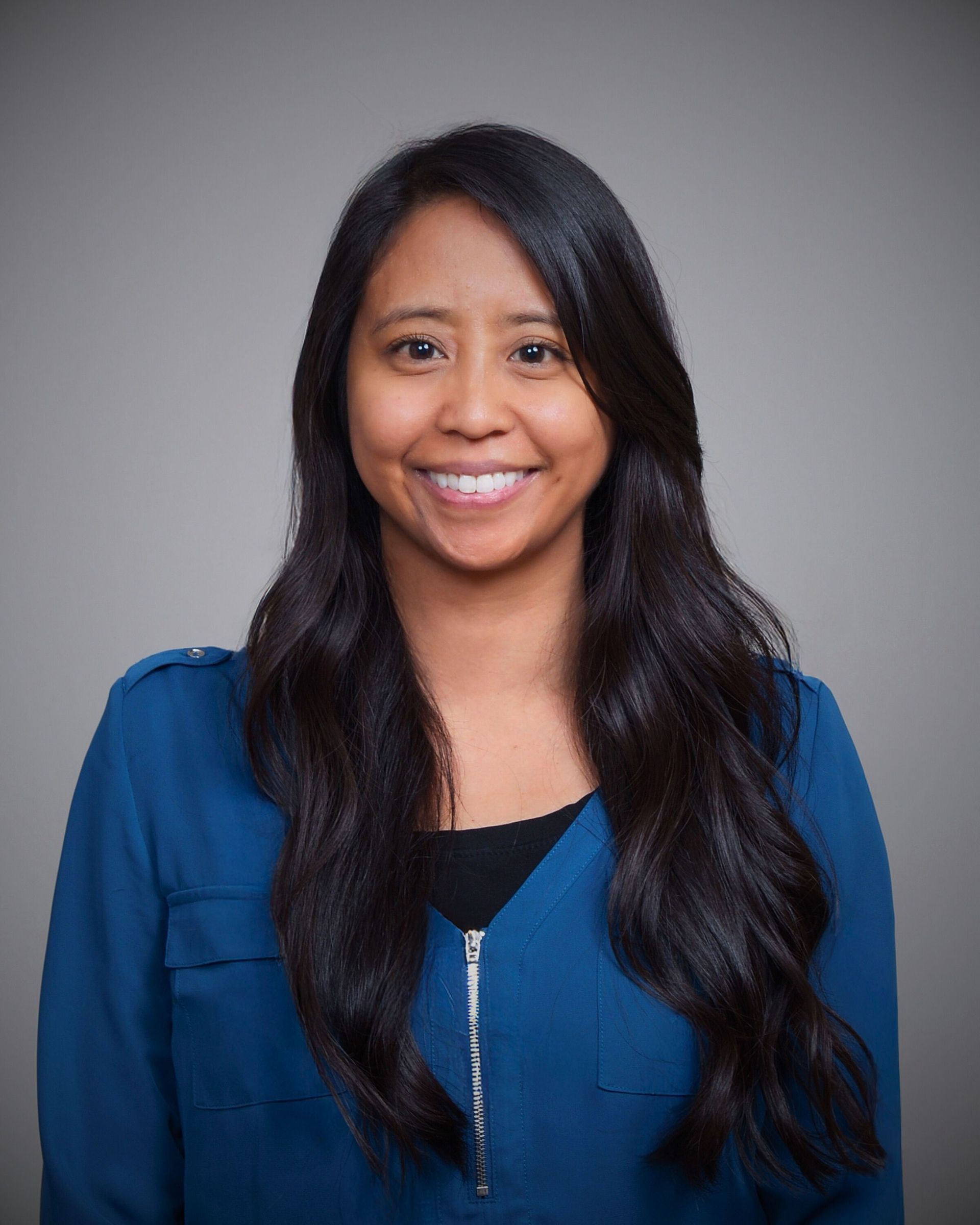 A woman in a dark blue shirt is smiling in front of a gray background.