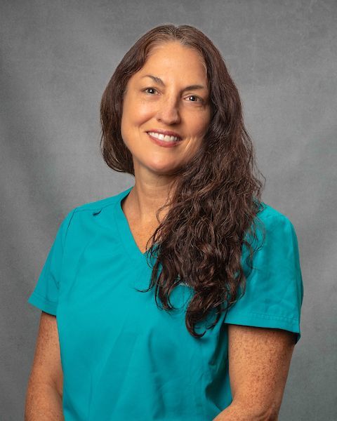 A woman wearing blue scrubs in front of a gray background.