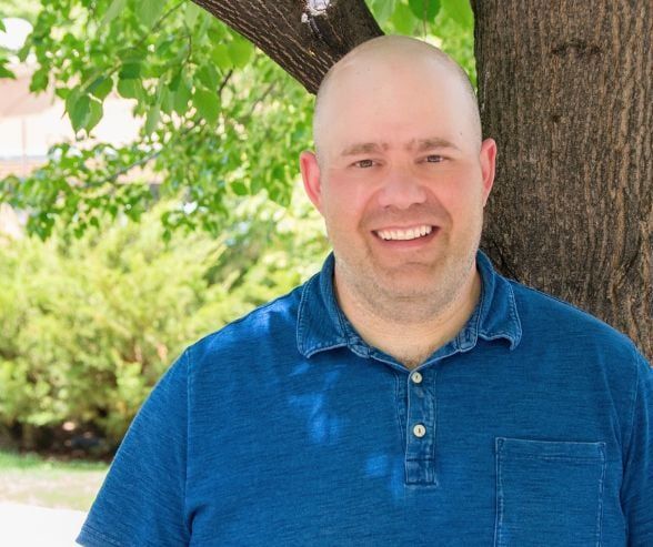 A bald man in a blue shirt is smiling while leaning against a tree.