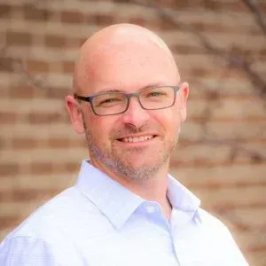 A bald man with glasses and a beard is smiling in front of a brick wall.