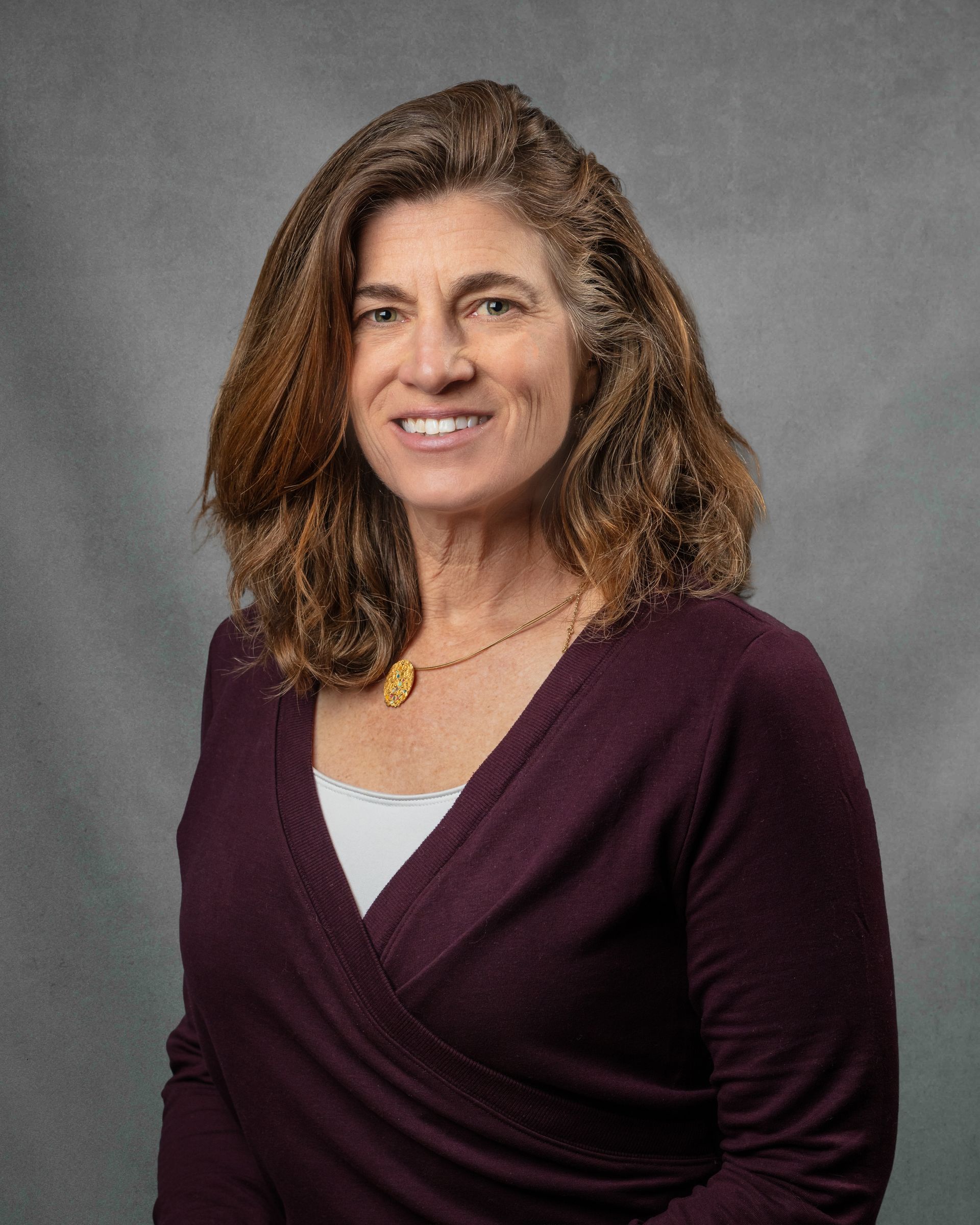 A woman in a dark purple shirt smiling in front of a gray background