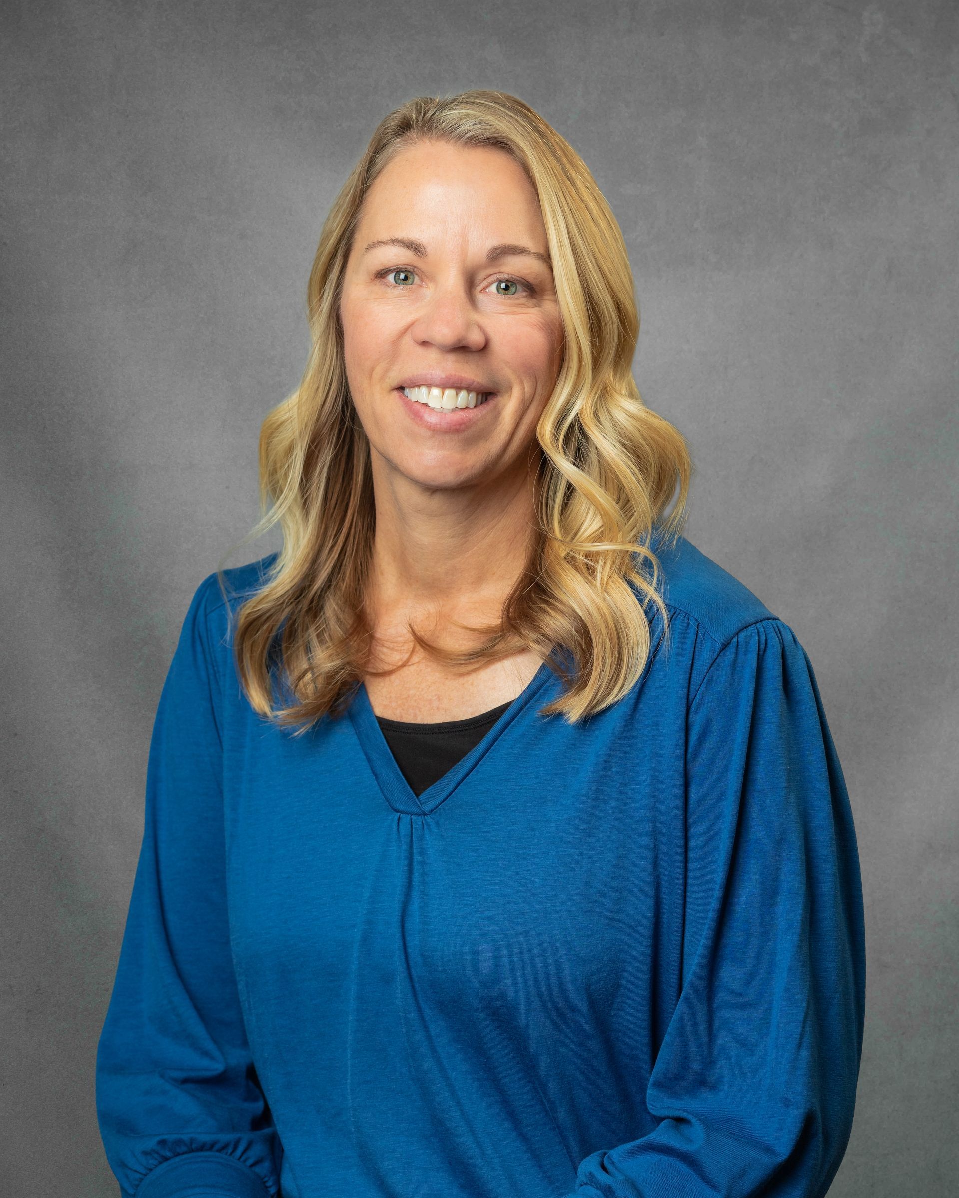 A woman in a blue shirt is smiling in front of a gray background.