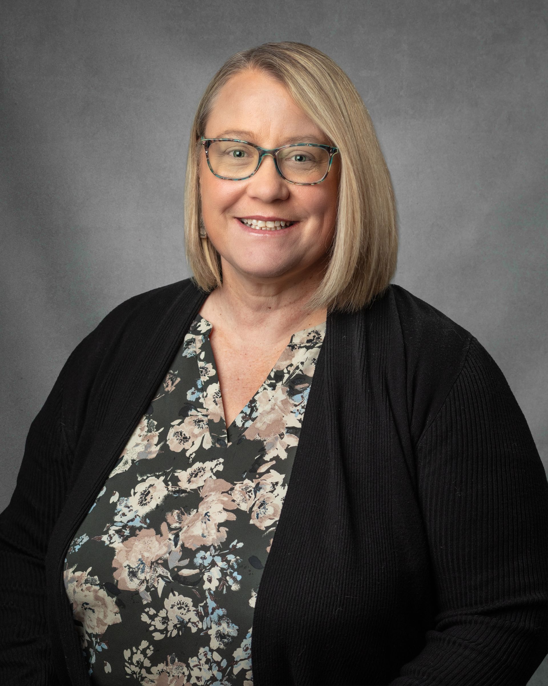 A woman wearing glasses and a black and white floral shirt with a black cardiganis smiling for the camera.
