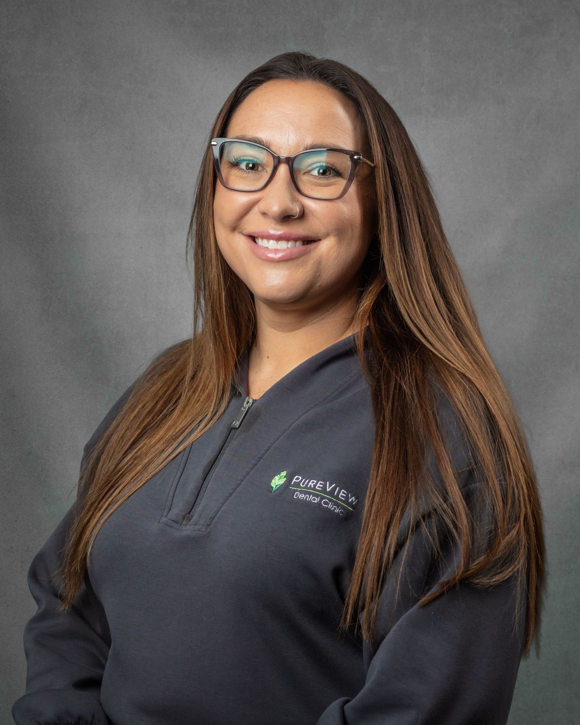 A woman wearing glasses and a gray shirt is smiling in front of a gray background.
