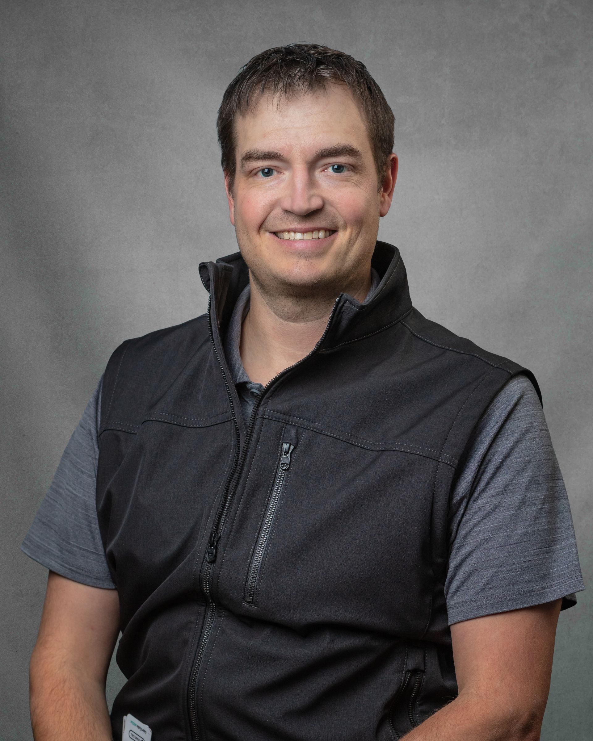 A man in a gray shirt and black vest is smiling.