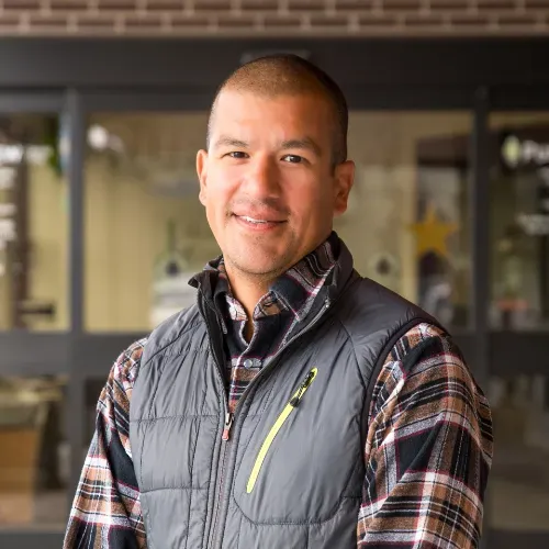 A man wearing a plaid shirt and a gray vest is smiling for the camera.