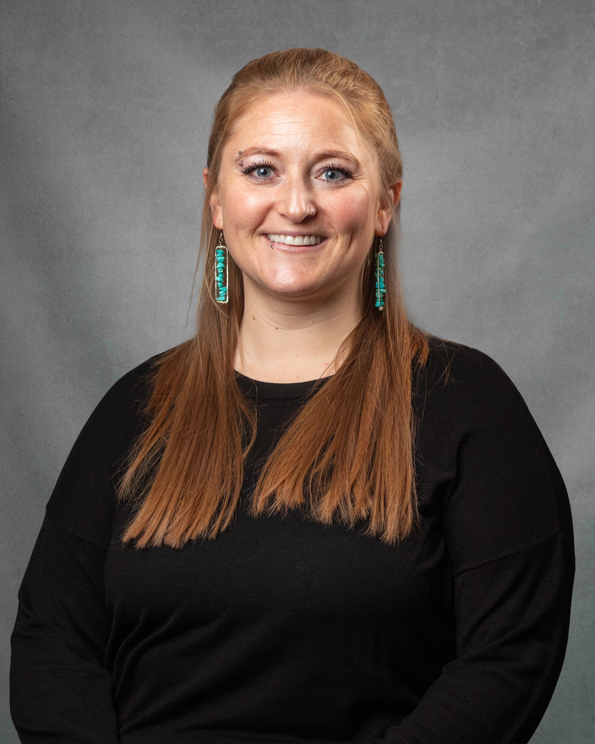 A woman with dangling earrings and an eyebrow piercing with a black shirt is smiling at the camera.