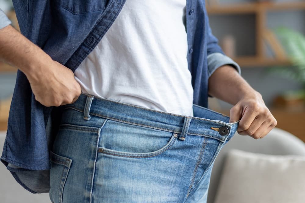 Man pulling out the waistband of oversized blue jeans to show successful weight loss results.