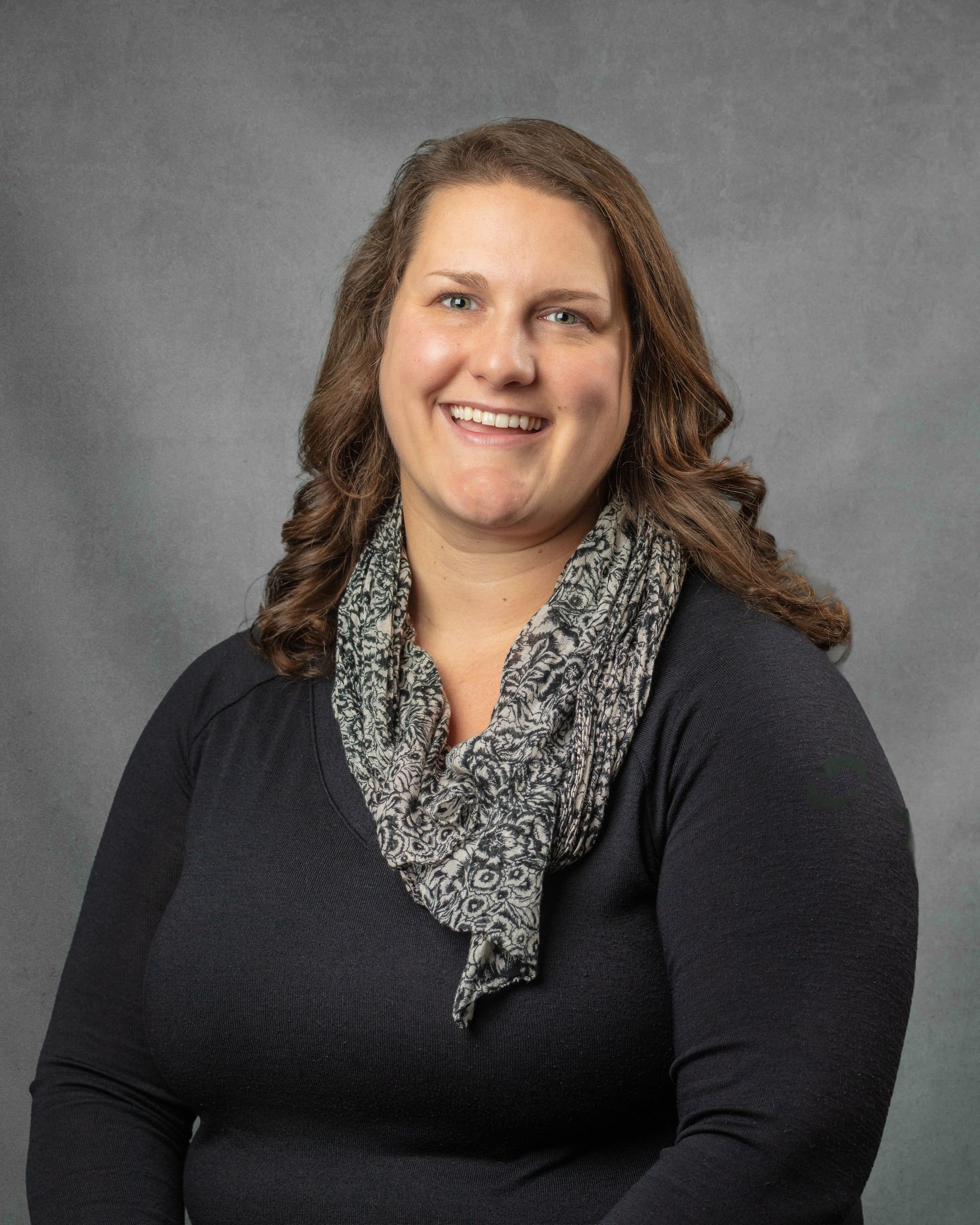 A woman with a black shirt and a gray scarf is smiling in front of a gray background.