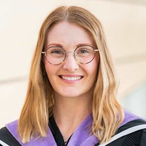 A woman wearing glasses and a graduation cap and gown is smiling for the camera.