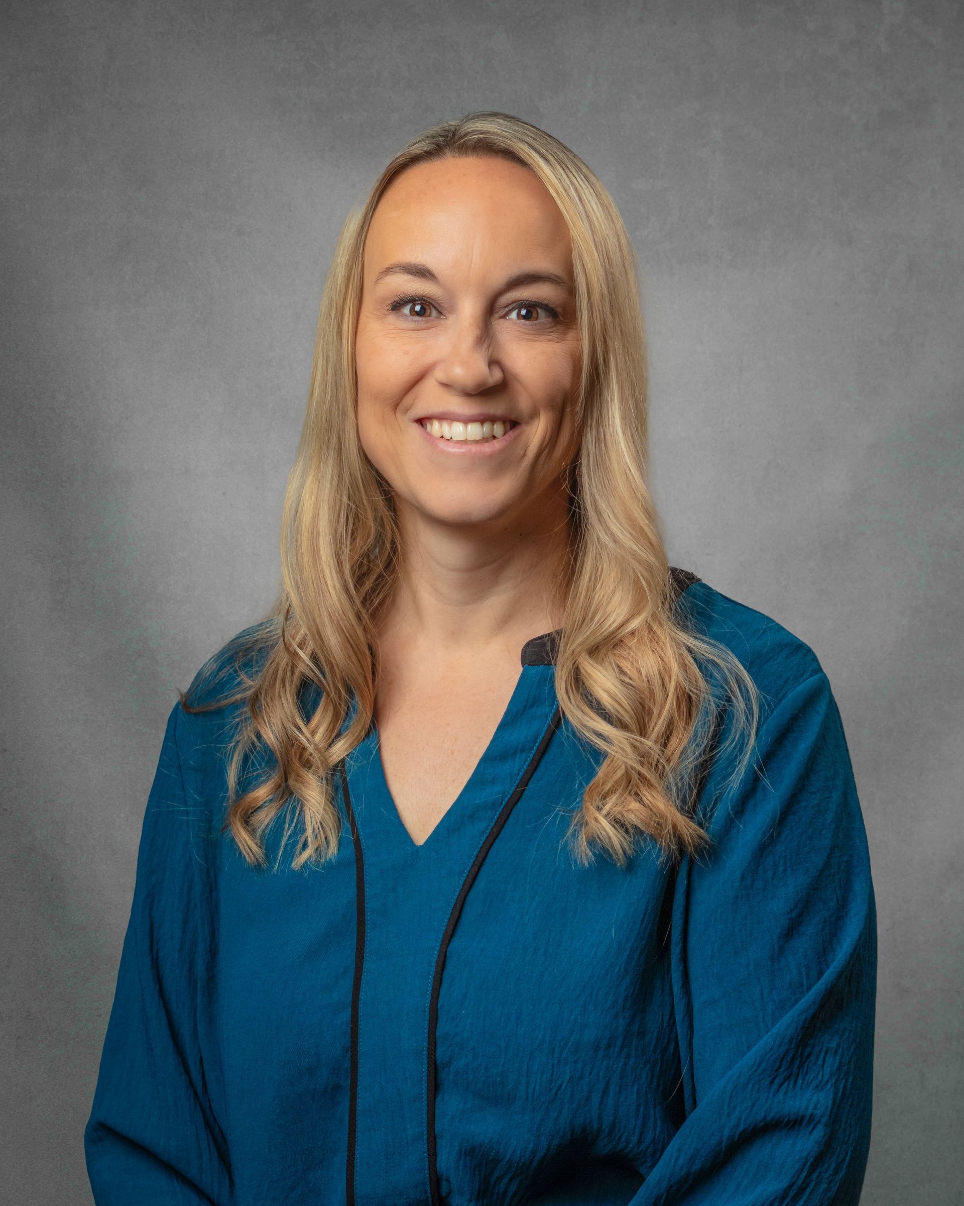 A woman in a blue shirt smiling in front of a gray background.