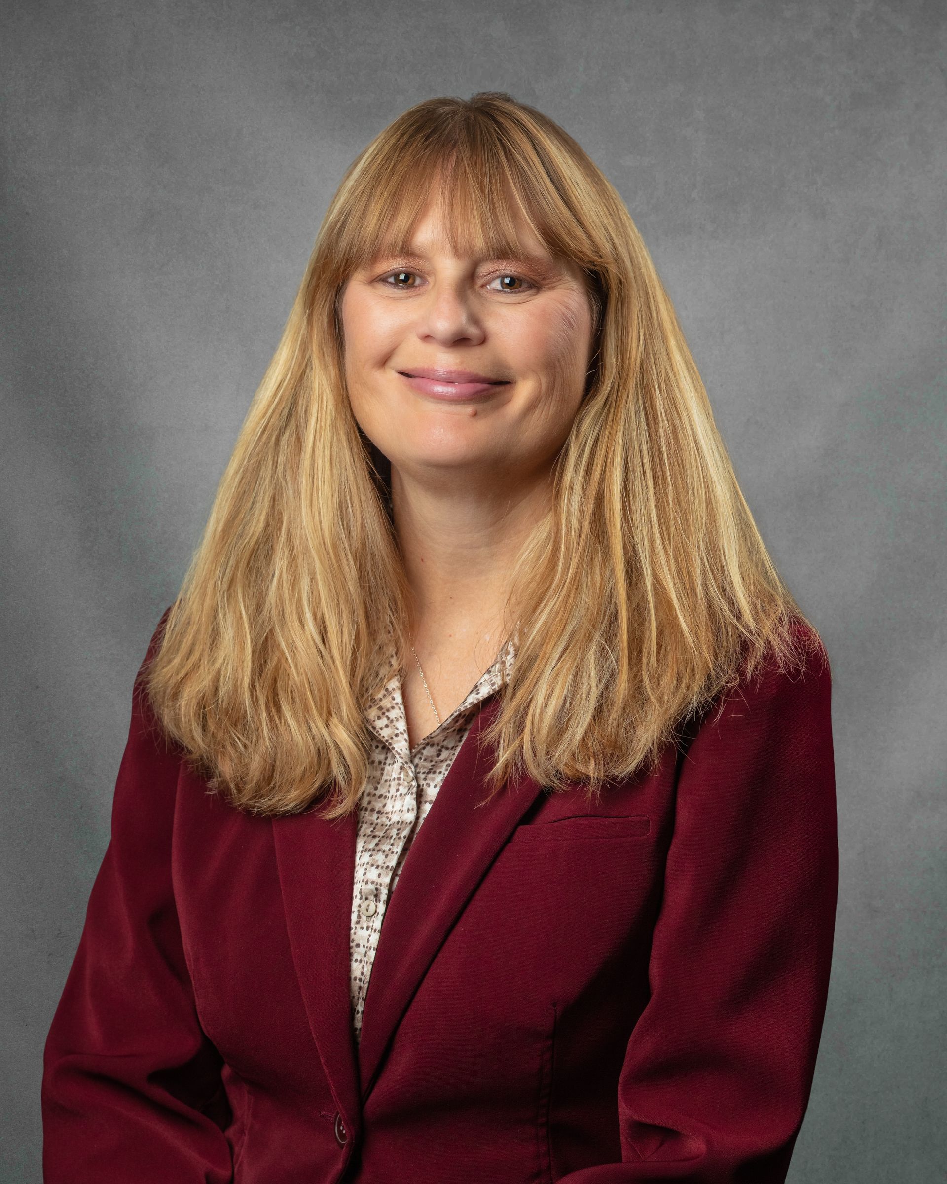 A woman wearing a red blazer smiling in front of a gray background.