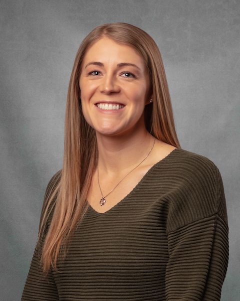 A woman in a olive green sweater is smiling in front of a gray background.