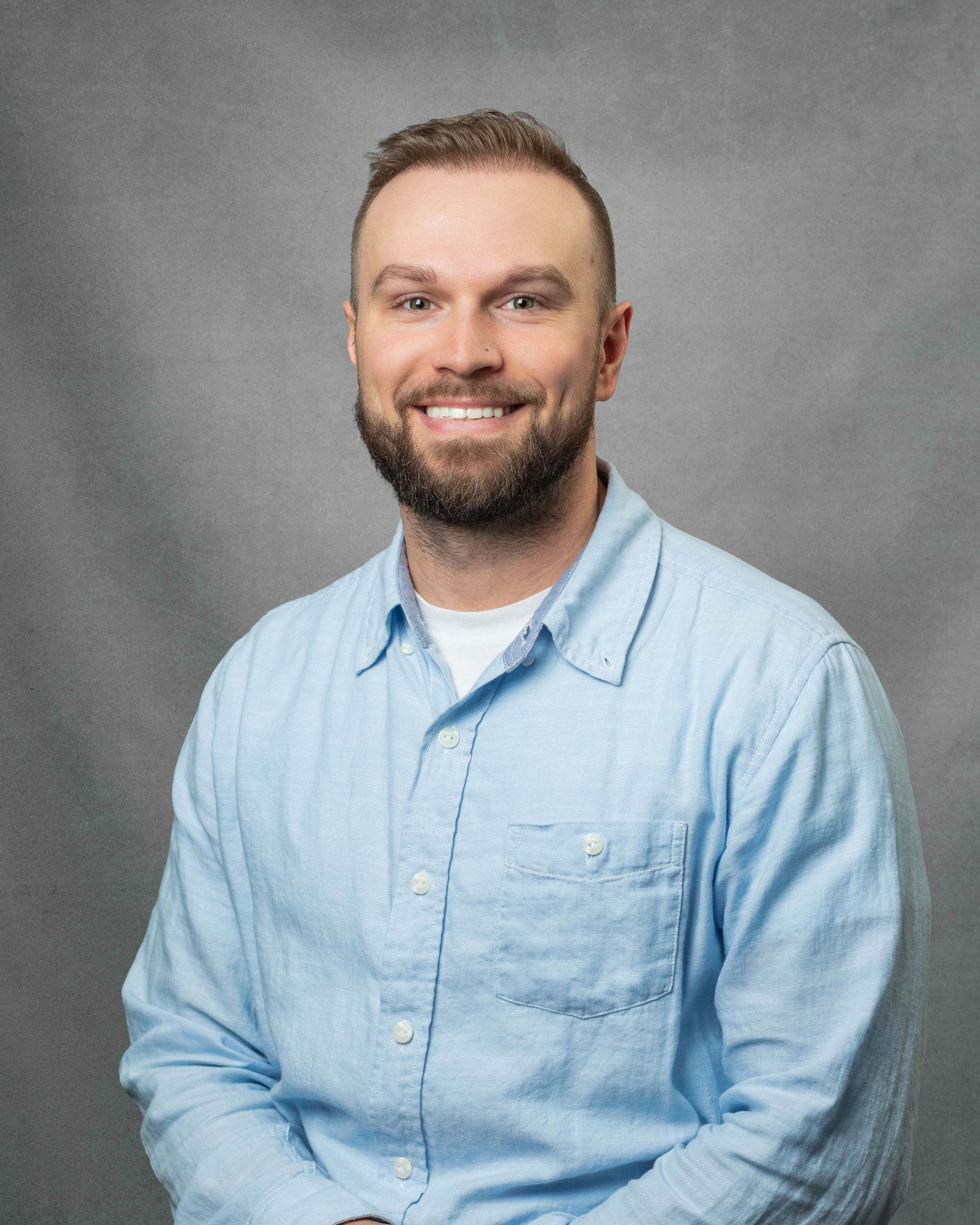 A man with a beard in a blue button down is smiling for the camera.