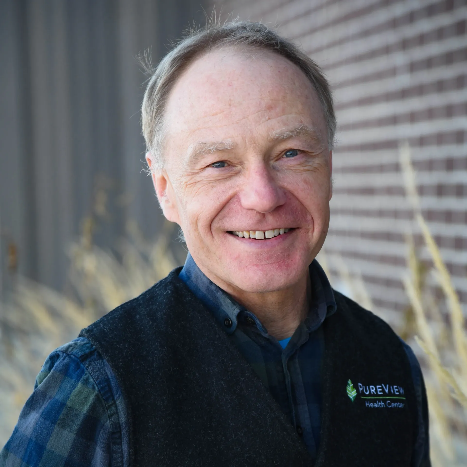 A man wearing a vest and a plaid shirt is smiling in front of a brick wall.