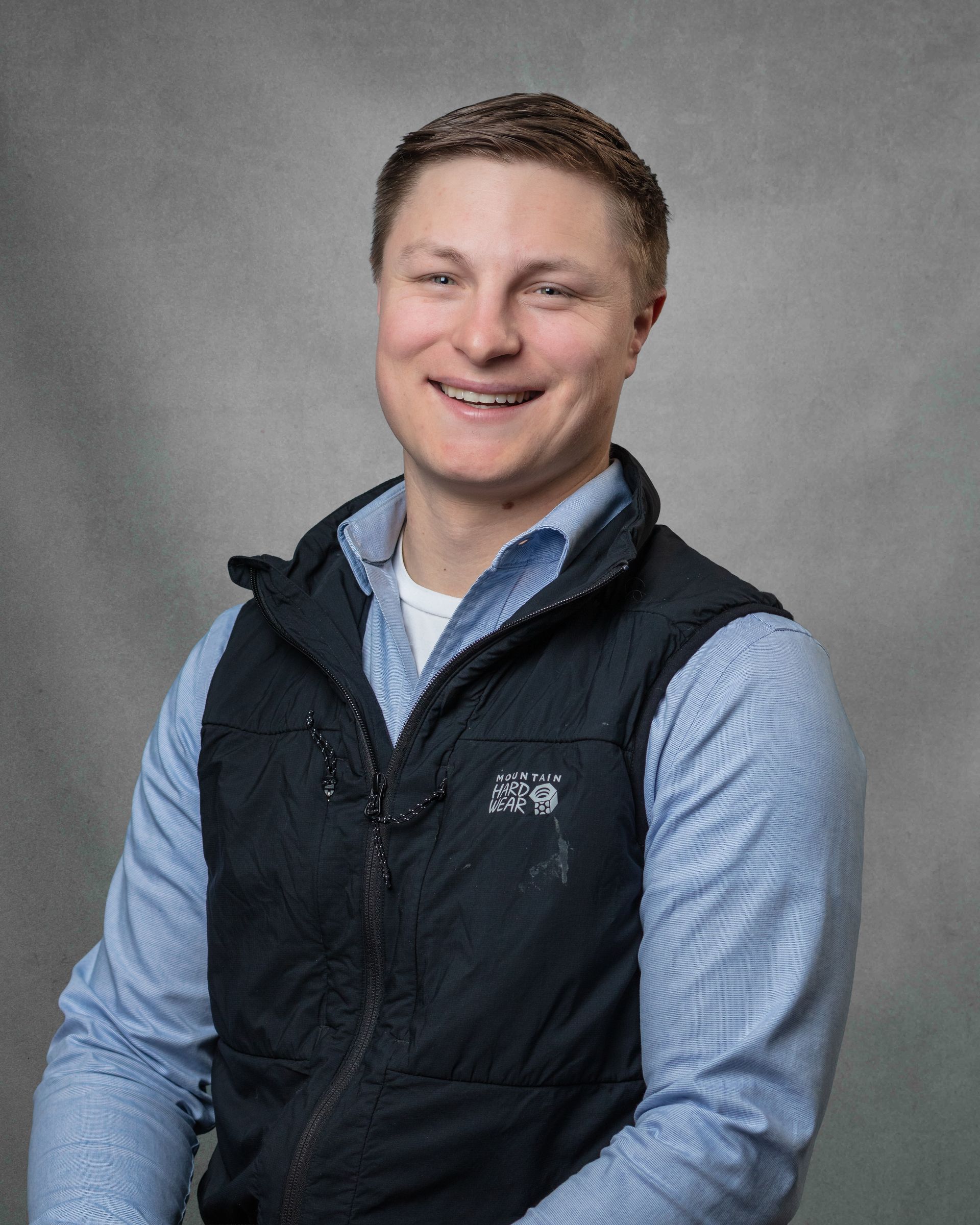 A man in a blue button down and a black vest is smiling for the camera.
