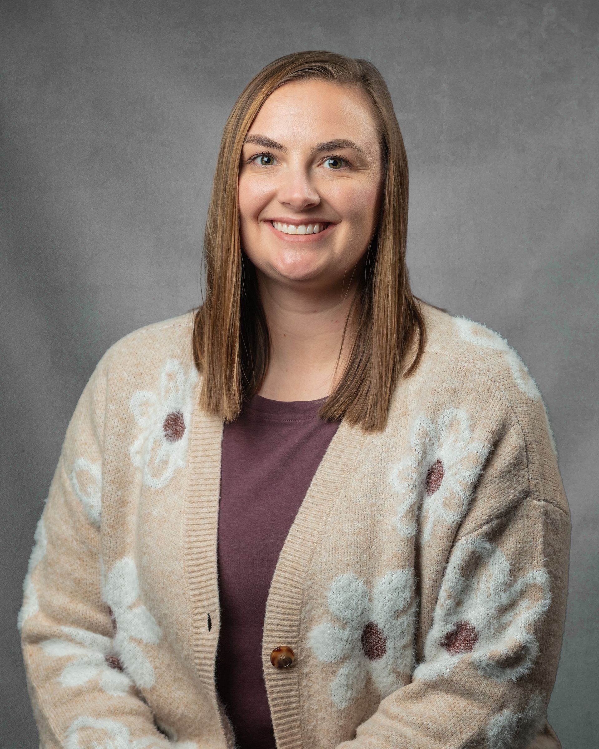 A woman in a light brown floral cardigan is smiling in front of a gray background