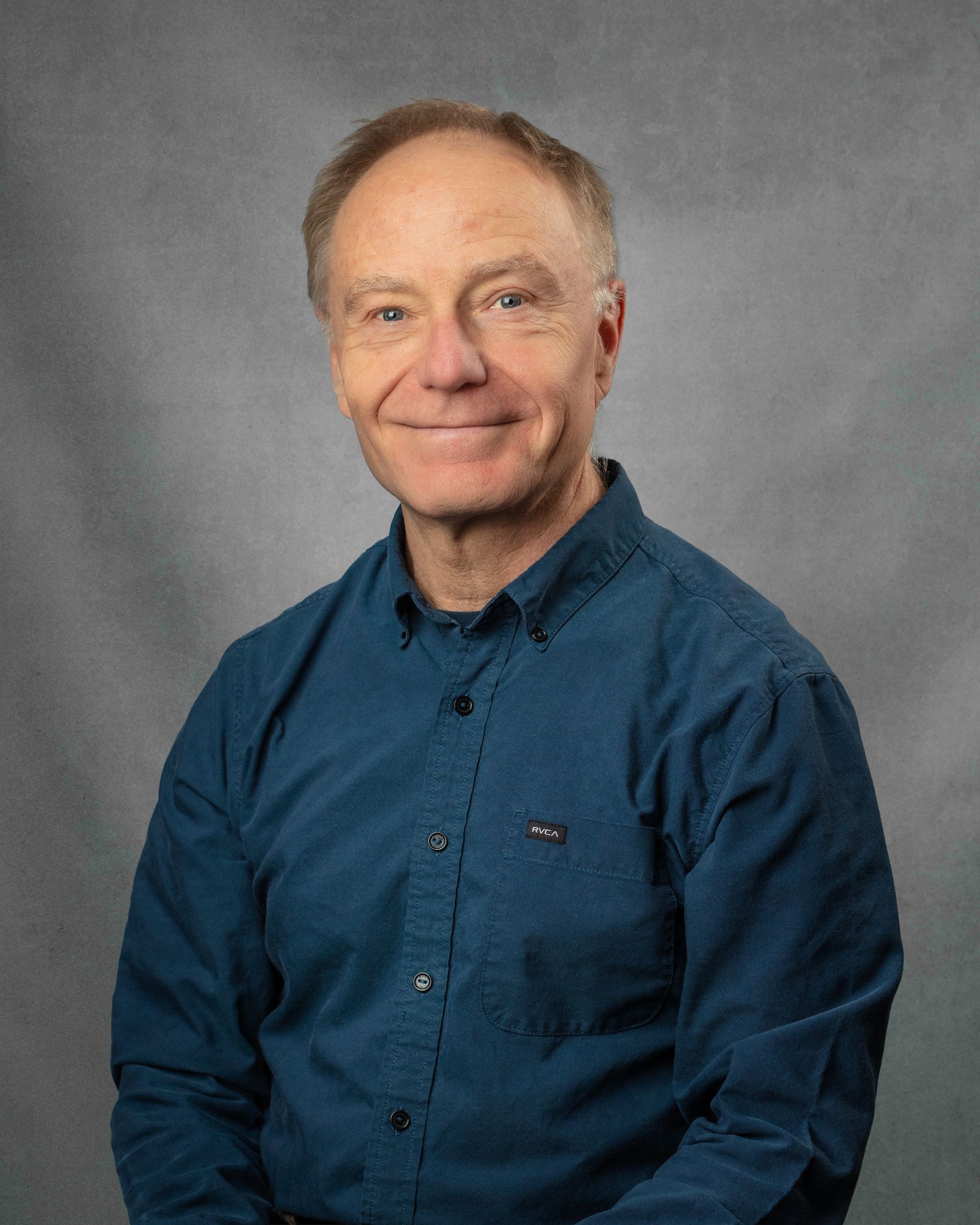 A man wearing a dark blue button down is smiling in front of a gray background.