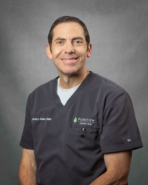 A man in a scrub top is smiling in front a gray background.