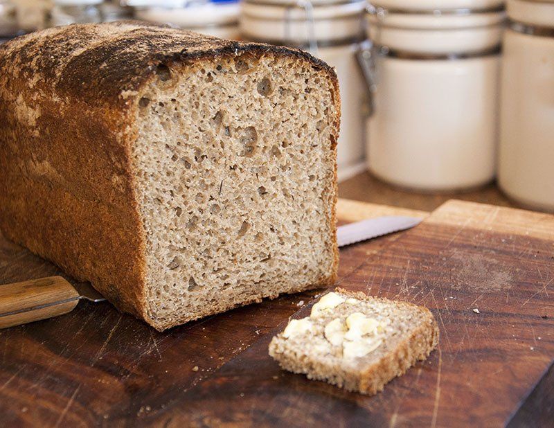 Loaf of bread with slice of buttered bread on a wooden board. Jars in background.