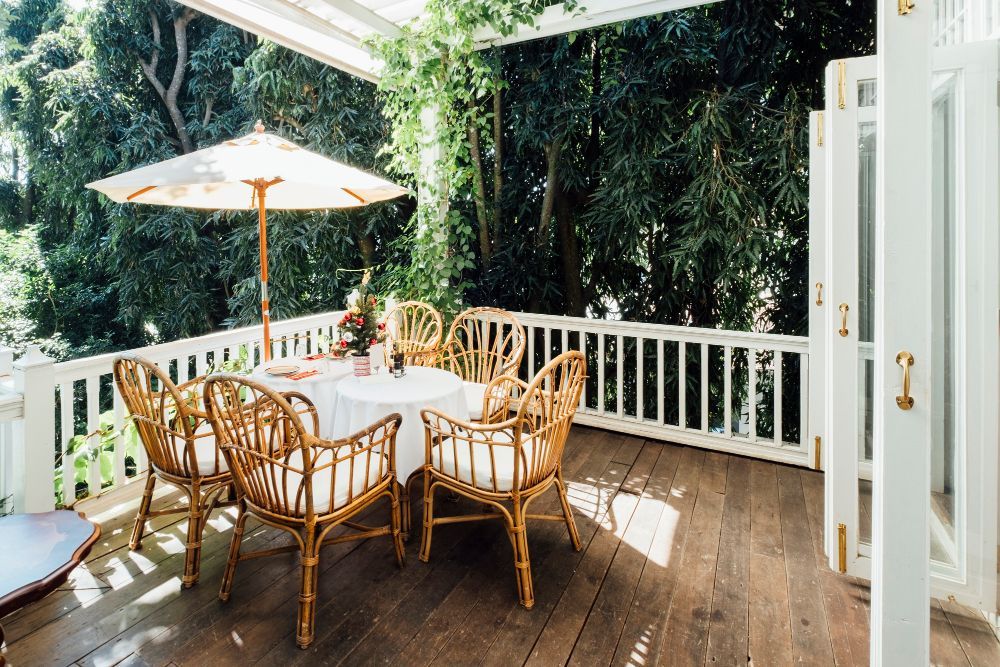 A white porch with a table and chairs shaded by an umbrella; lush greenery in the background.