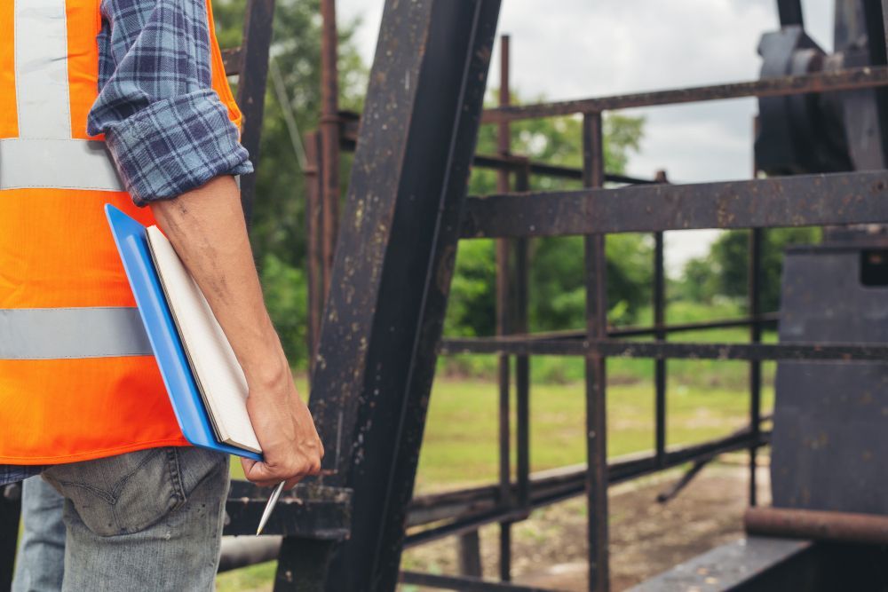 Person in orange vest holds a clipboard near industrial equipment outdoors.