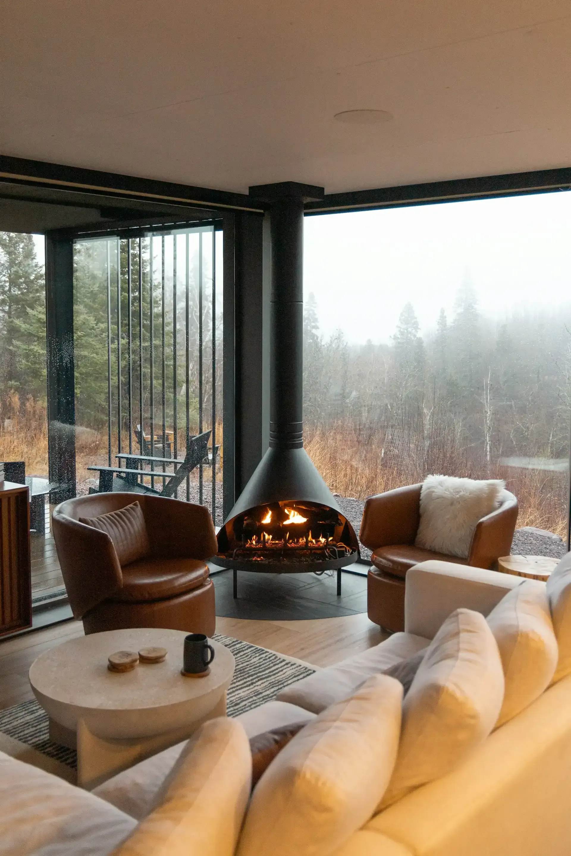 Cozy living room with fireplace, leather chairs, and large window overlooking a forest on a rainy day.