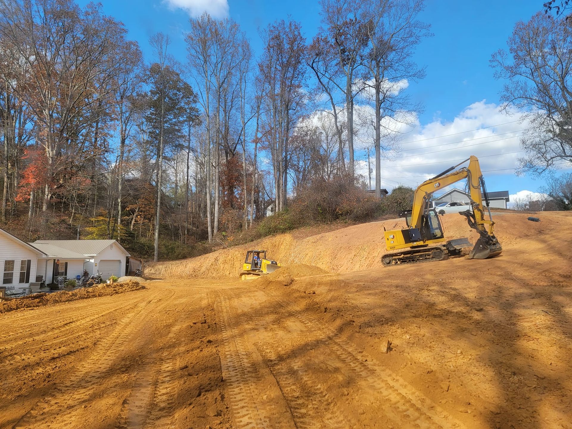 A yellow excavator is working on a dirt field in front of a house.