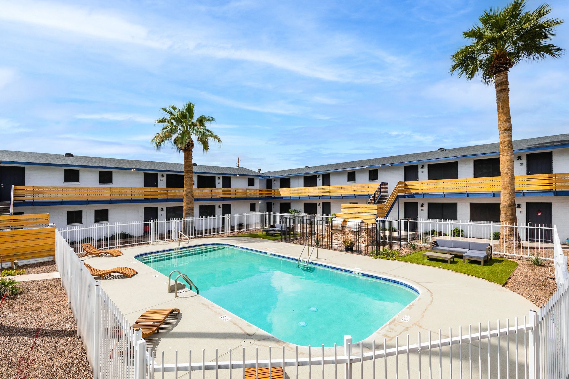 Apartment complex with a pool, palm trees, and white fencing. Blue sky.