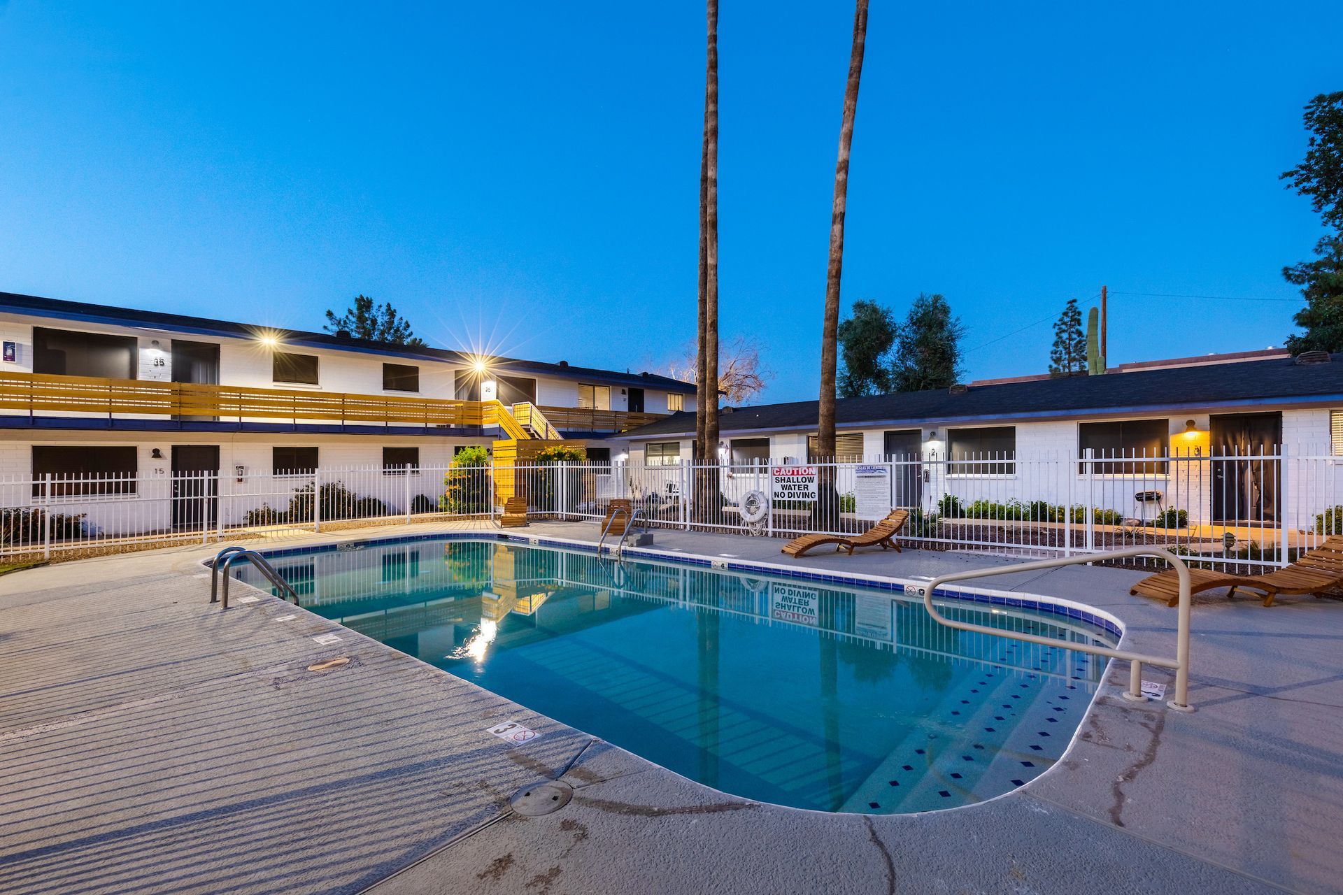 Swimming pool in front of a white motel at dusk, with palm trees and a clear blue sky.