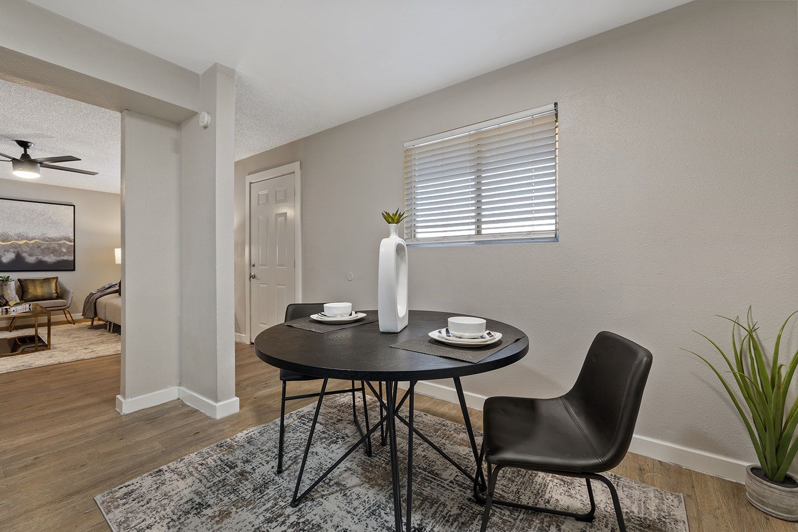 Dining area with round black table, two chairs, rug, window blinds, and open doorway to living room.