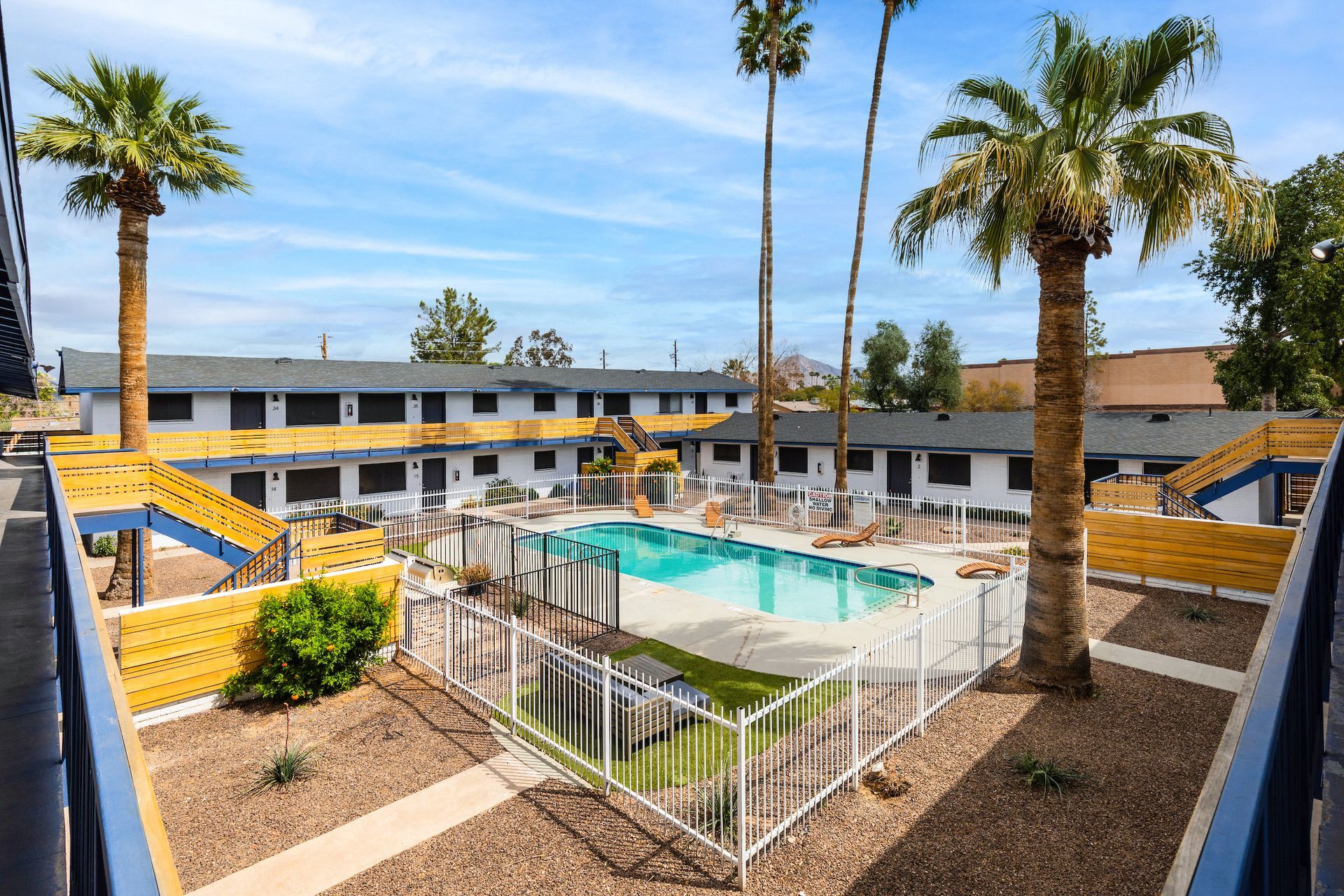Apartment complex with a pool, surrounded by palm trees and two-story buildings, bright sunny day.