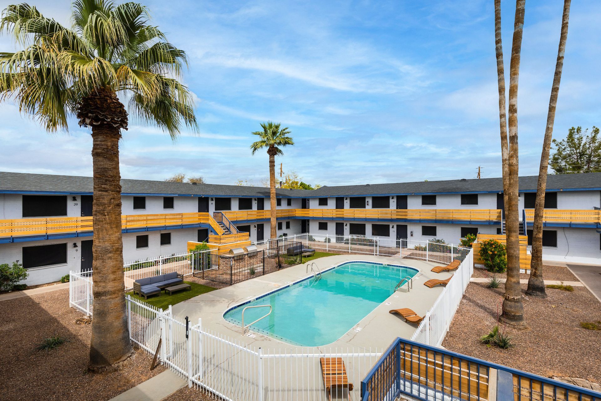 Apartment complex with a pool, palm trees, and two-story buildings, under a blue sky.
