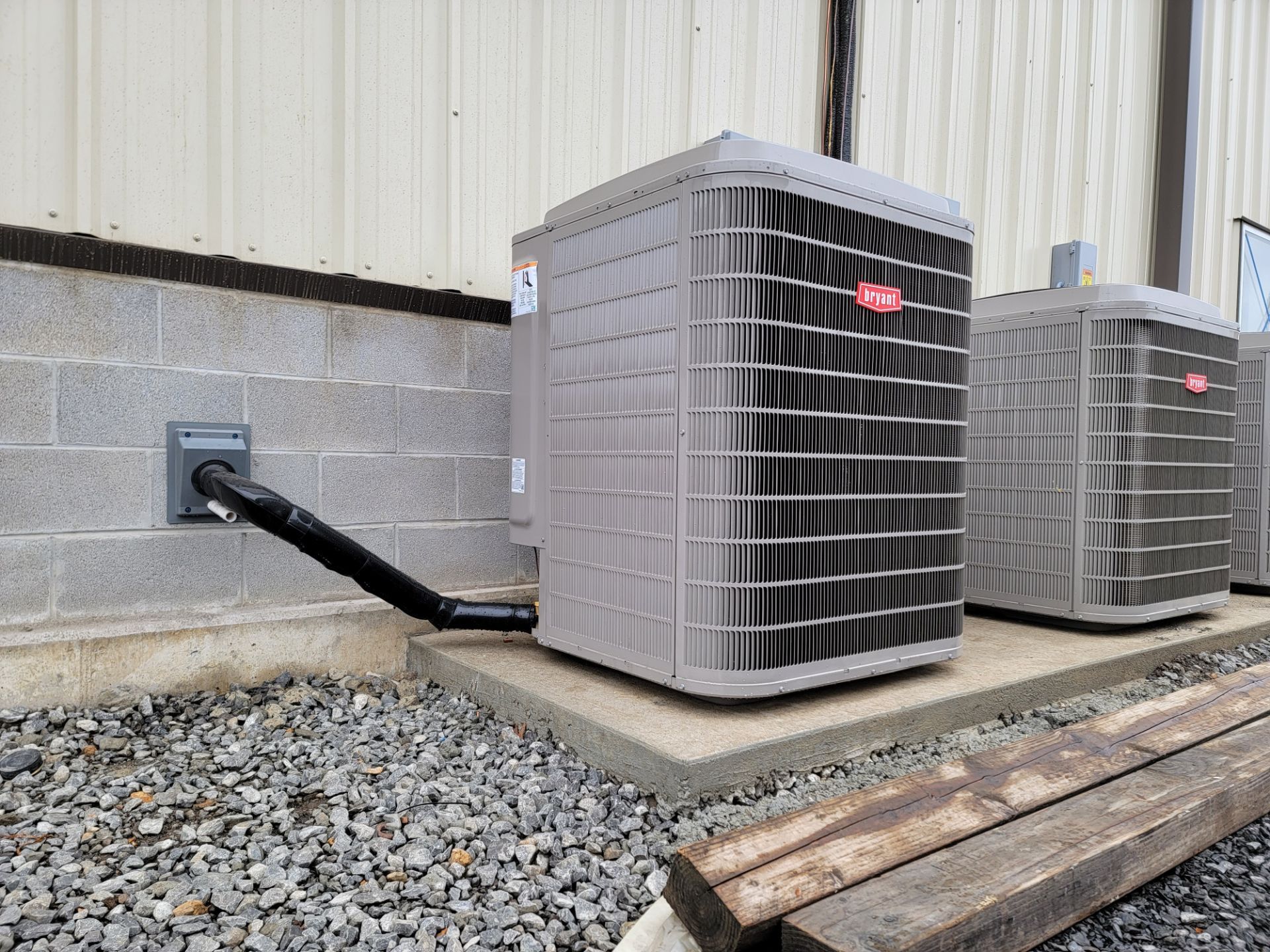 Two outdoor air conditioning units beside a building, connected by black conduit on a concrete pad.