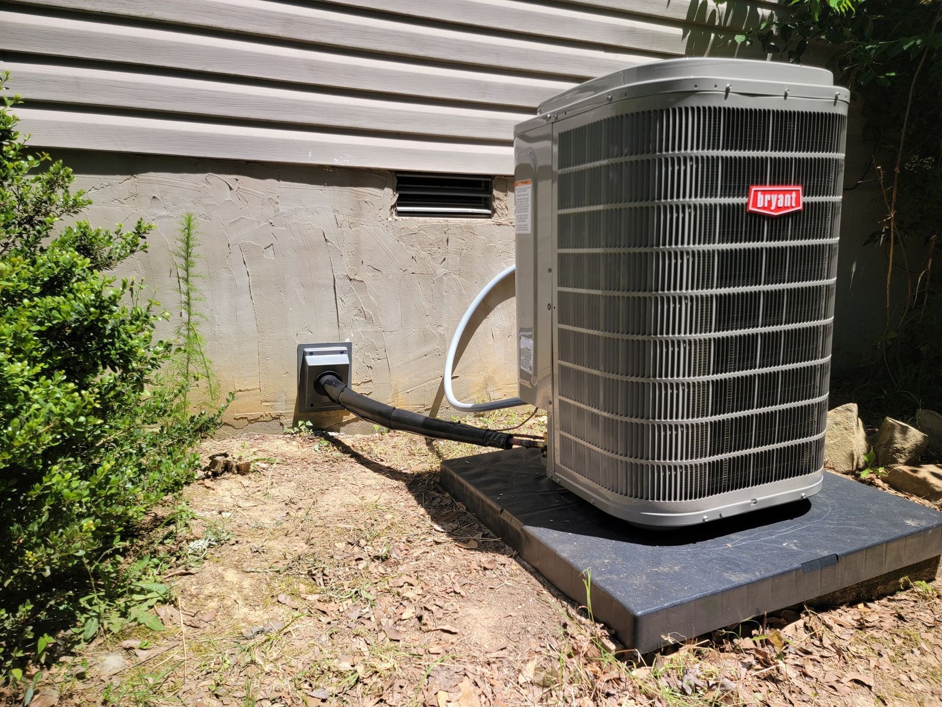 Outdoor HVAC unit beside a house, with a black pad and attached wiring near the wall.