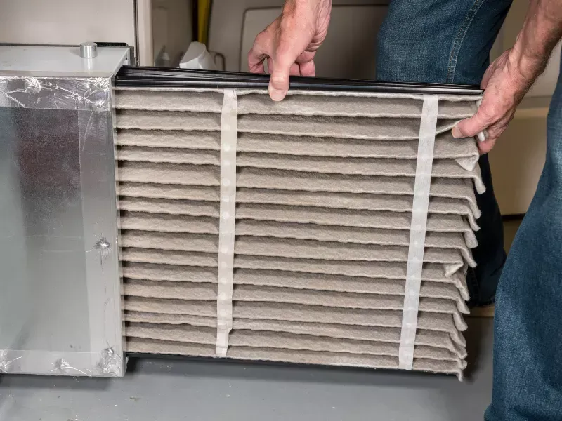 Hands holding a large pleated HVAC filter cartridge beside metal equipment
