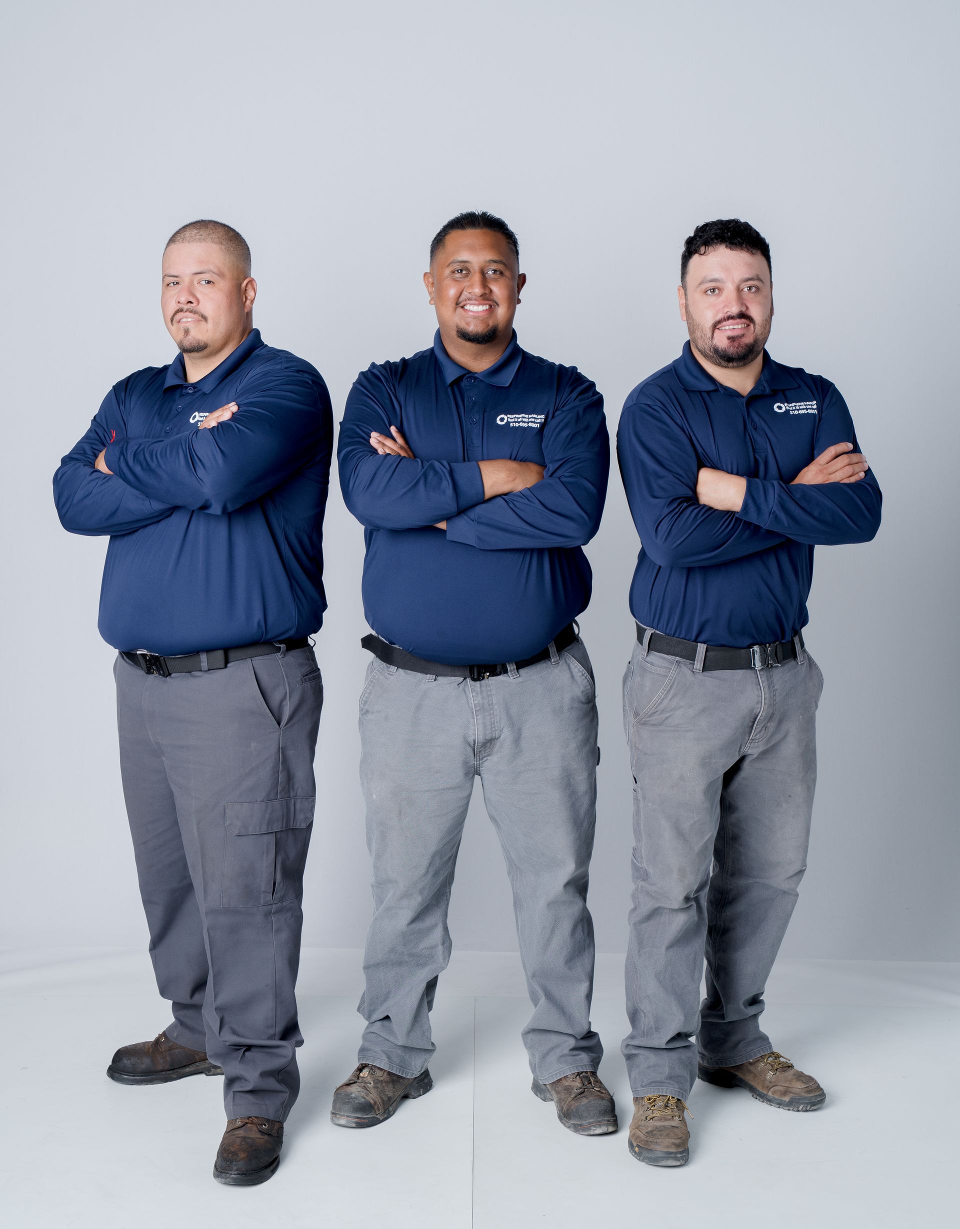 Three men stand confidently side by side, arms crossed, wearing matching navy blue shirts and gray pants
