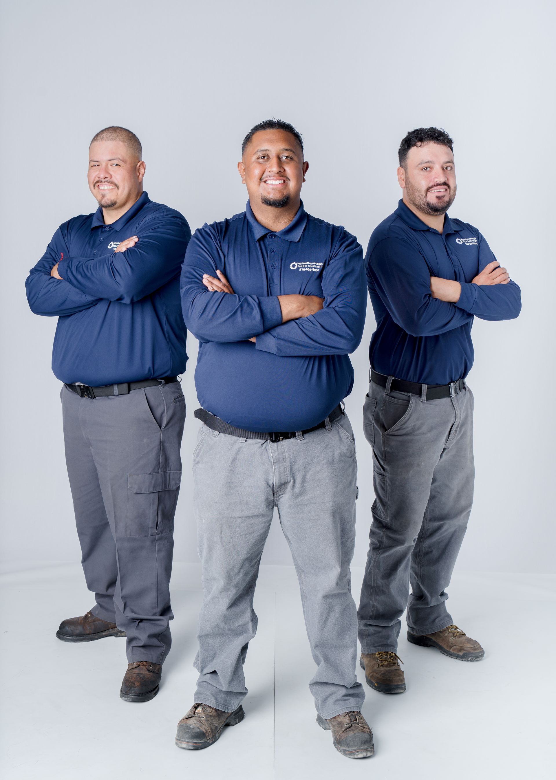 Three men stand confidently side by side, arms crossed, wearing matching navy blue shirts and gray pants