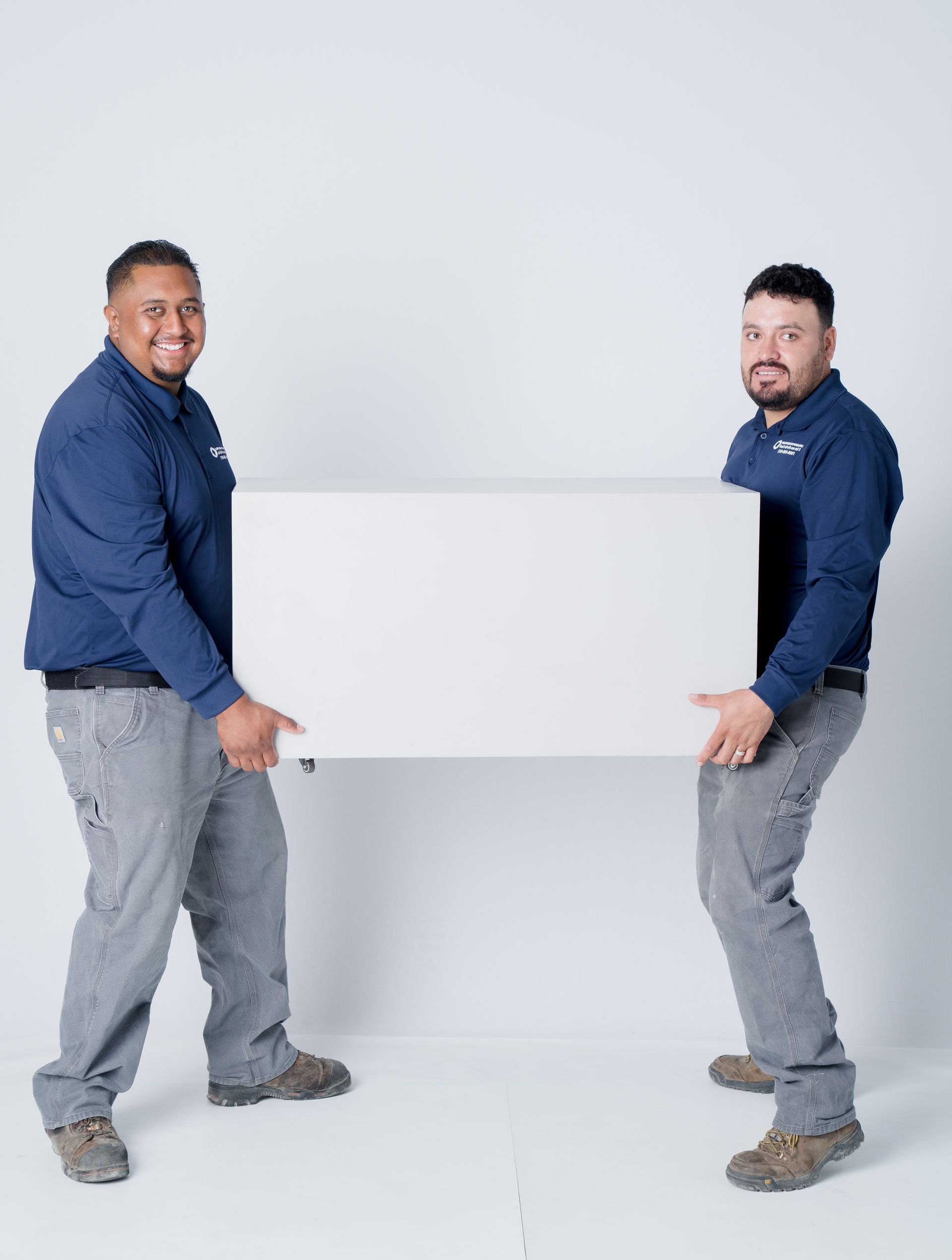 Two smiling men in matching navy shirts lift a white box.