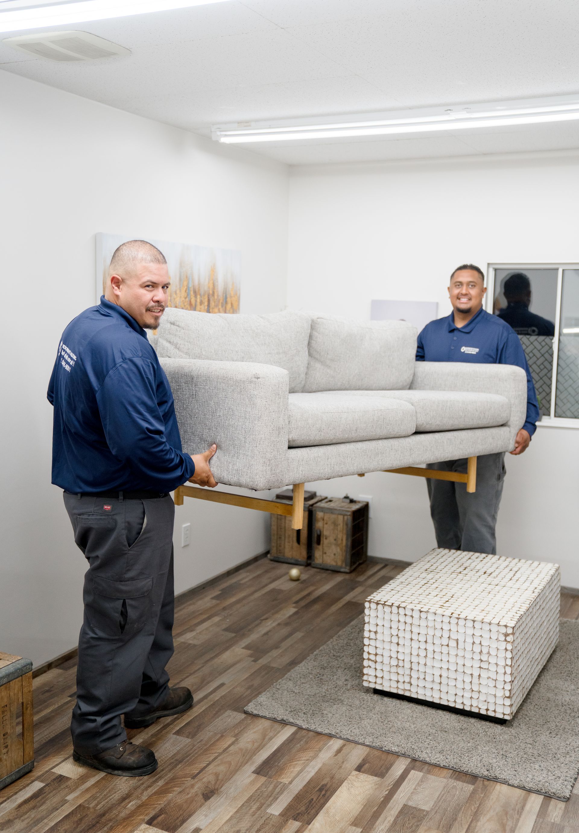 Two smiling men in matching navy shirts lift a gray sofa.
