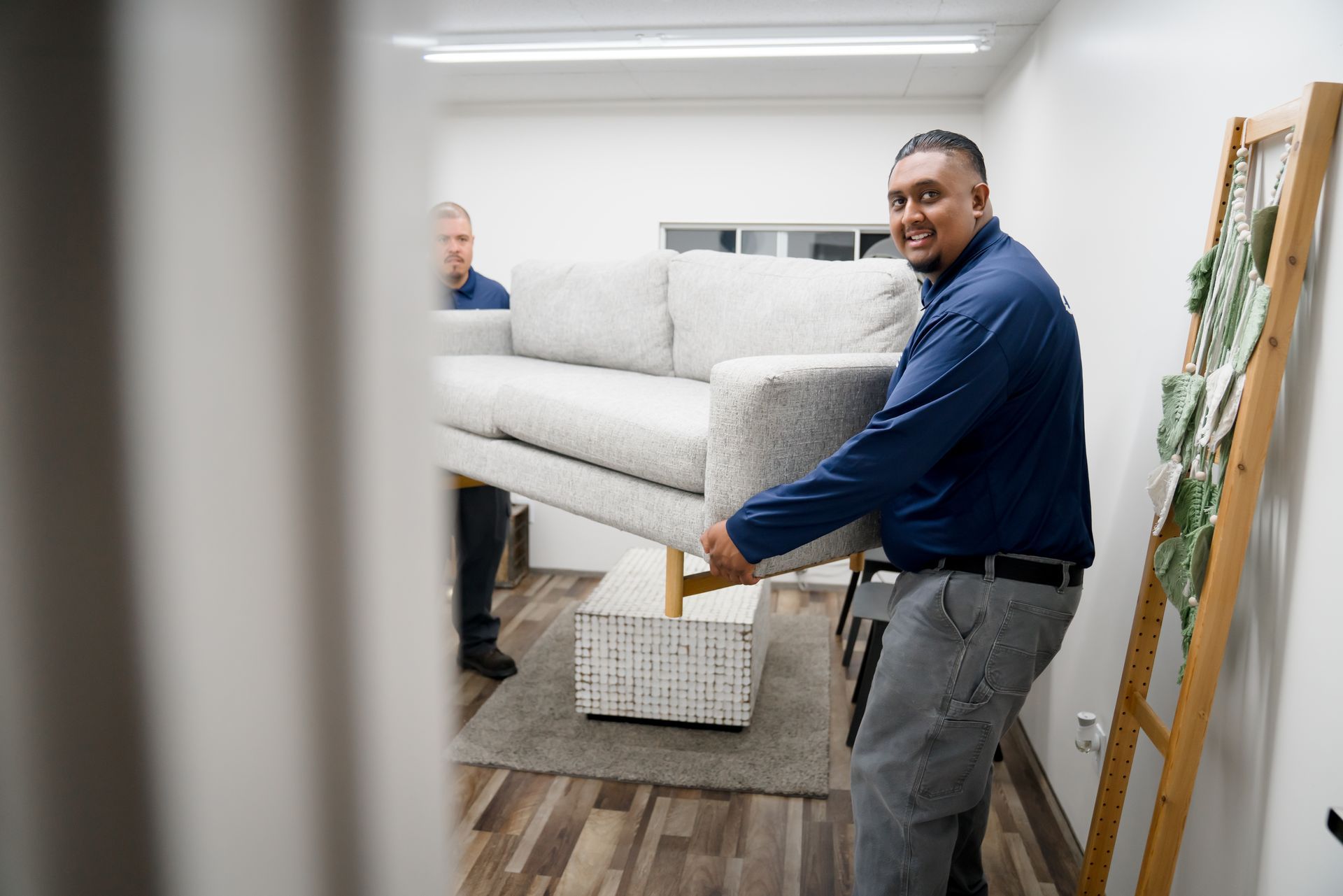 Two smiling men in matching navy shirts lift a gray sofa.