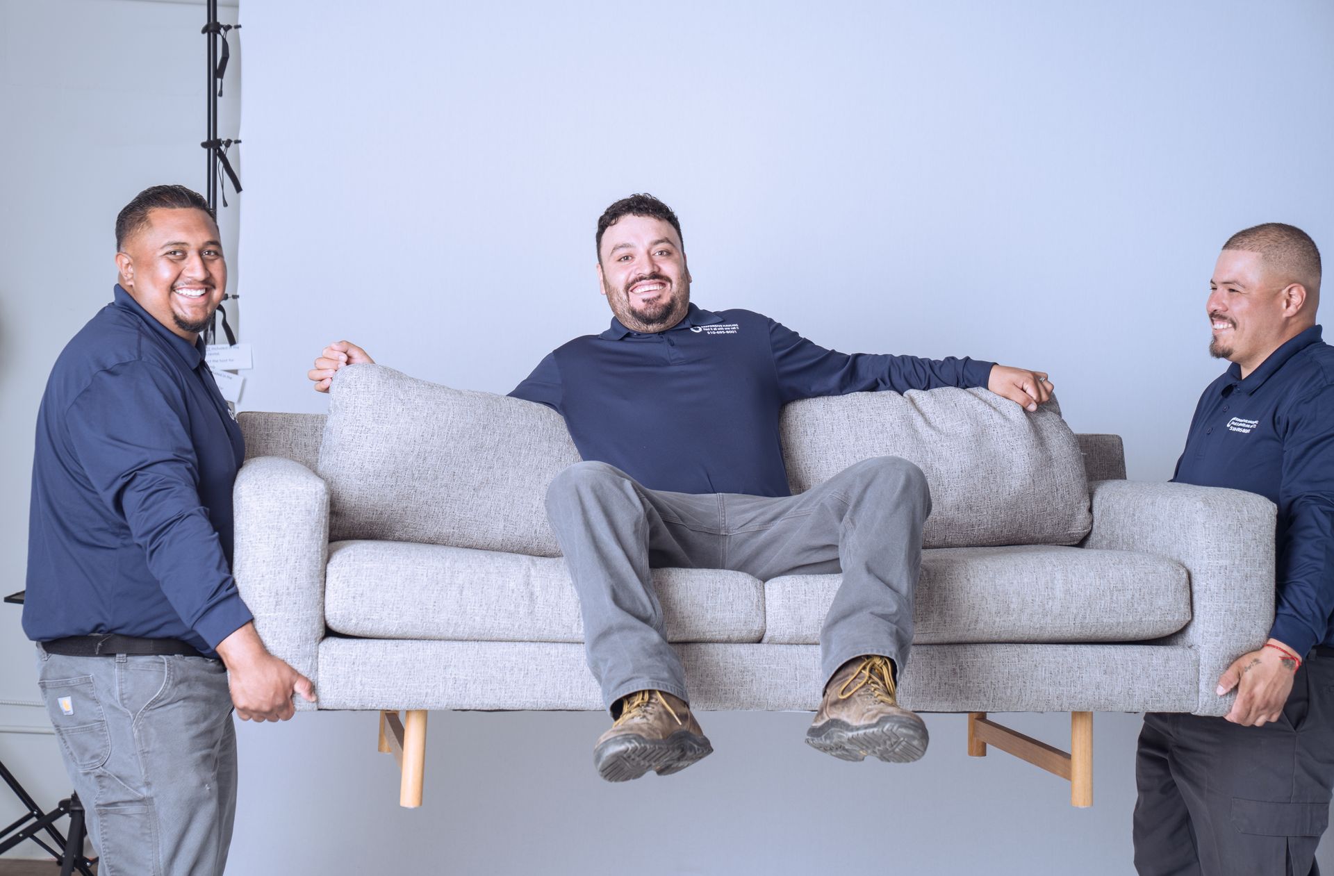 Two smiling men in matching navy shirts lift a gray sofa while a third man sits on the sofa.