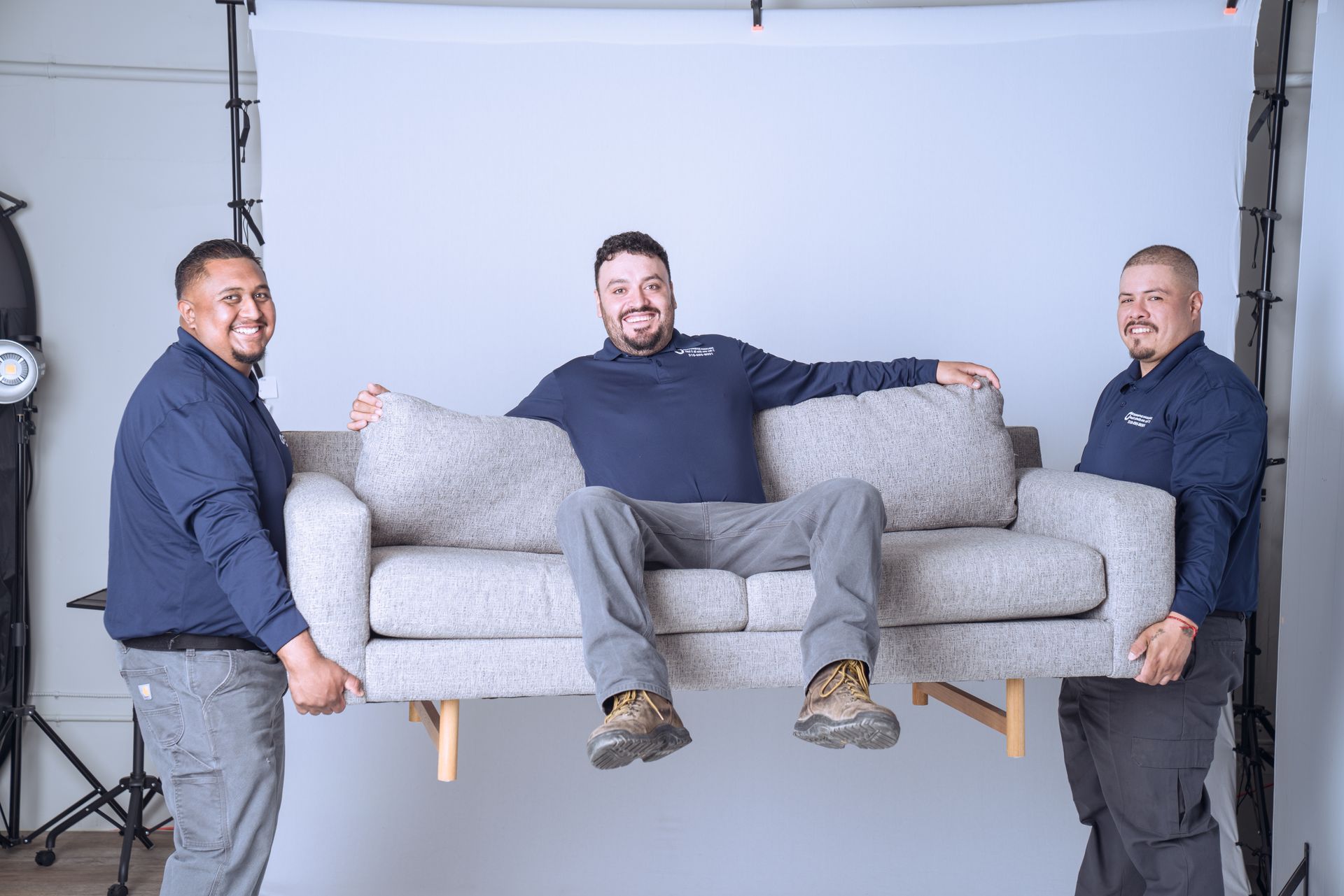 Two smiling men in matching navy shirts lift a gray sofa while a third man sits on the sofa.