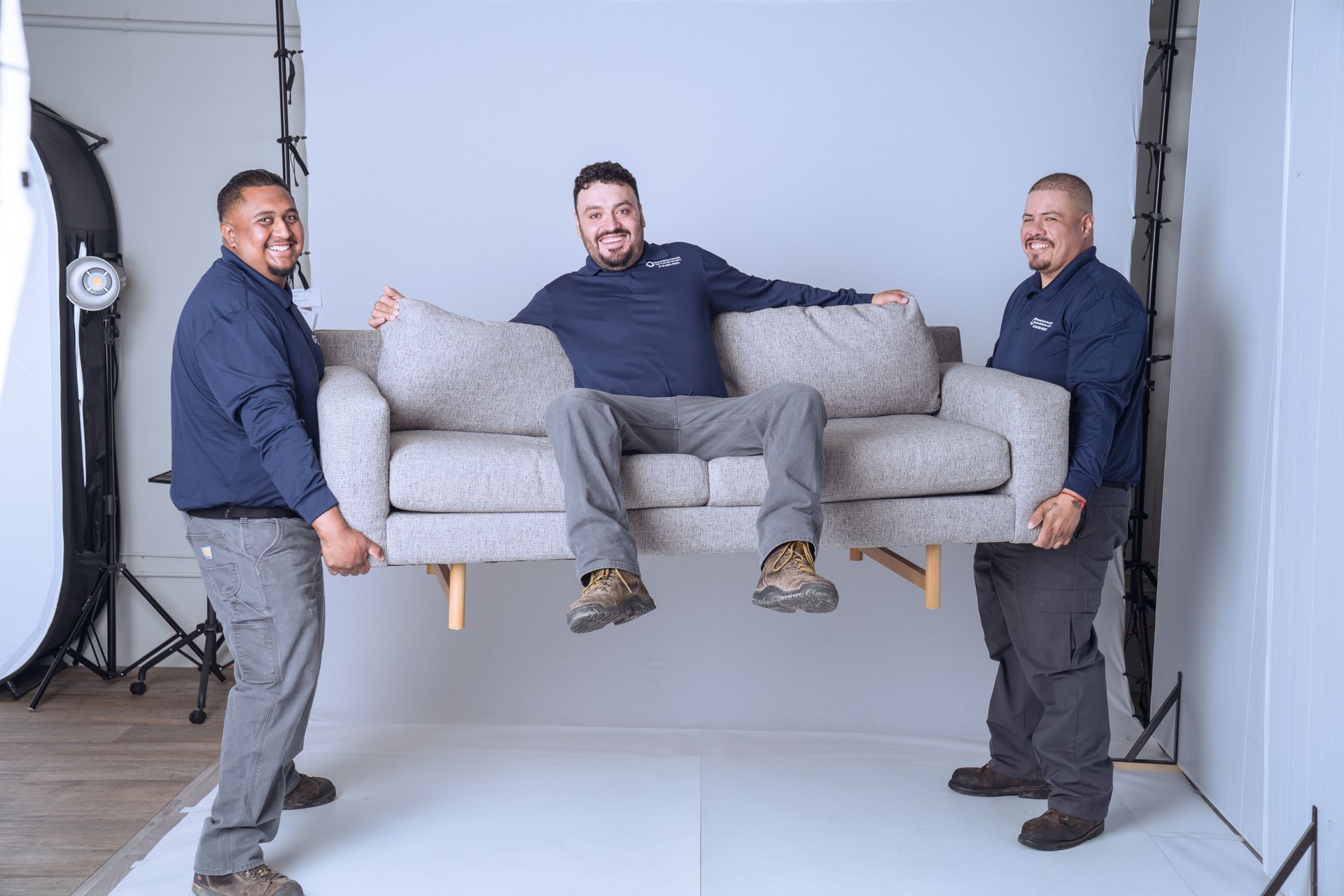 Two smiling men in matching navy shirts lift a gray sofa while a third man sits on the sofa.