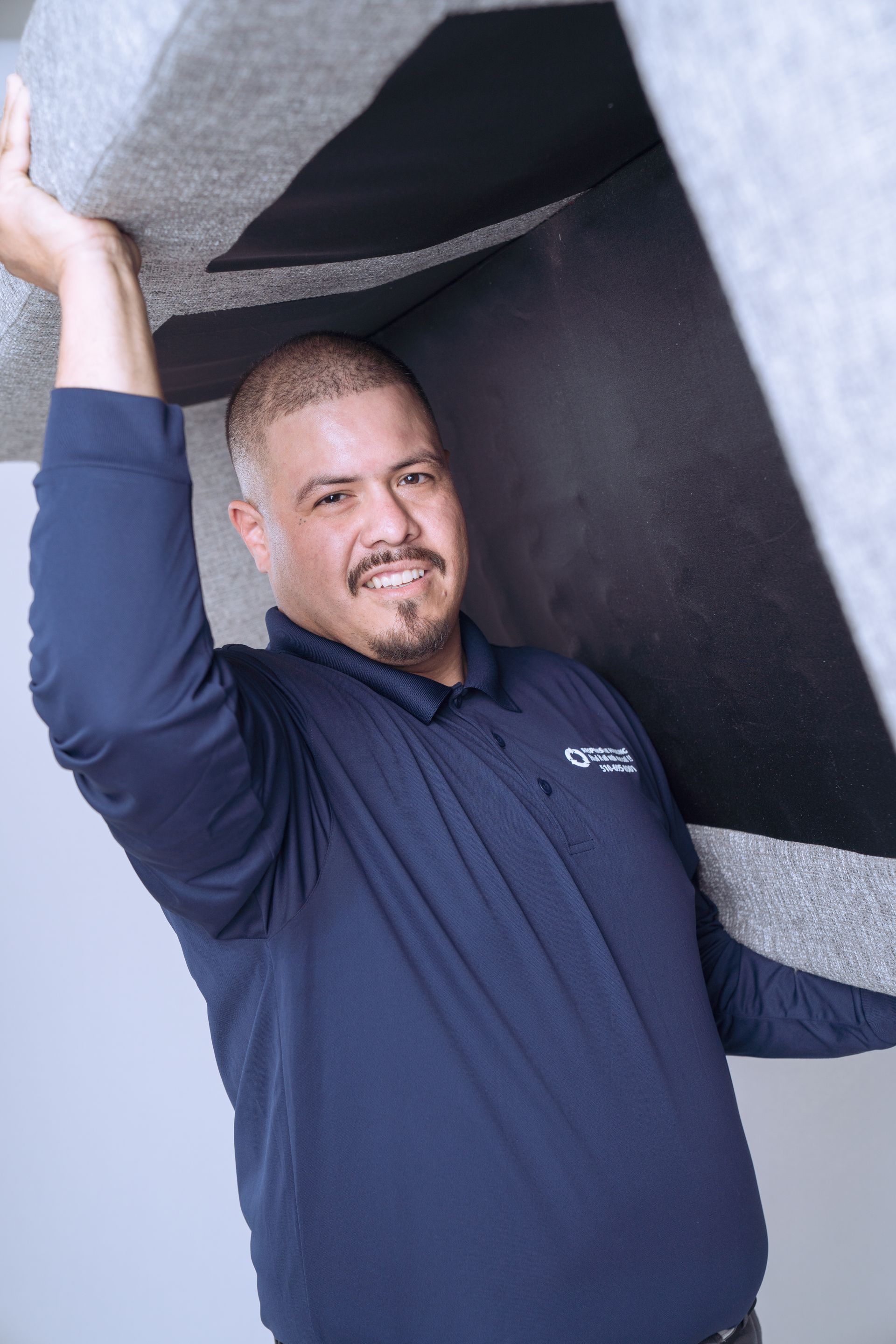 A man in a navy shirt and gray pants lifts a large beige sofa above his head.